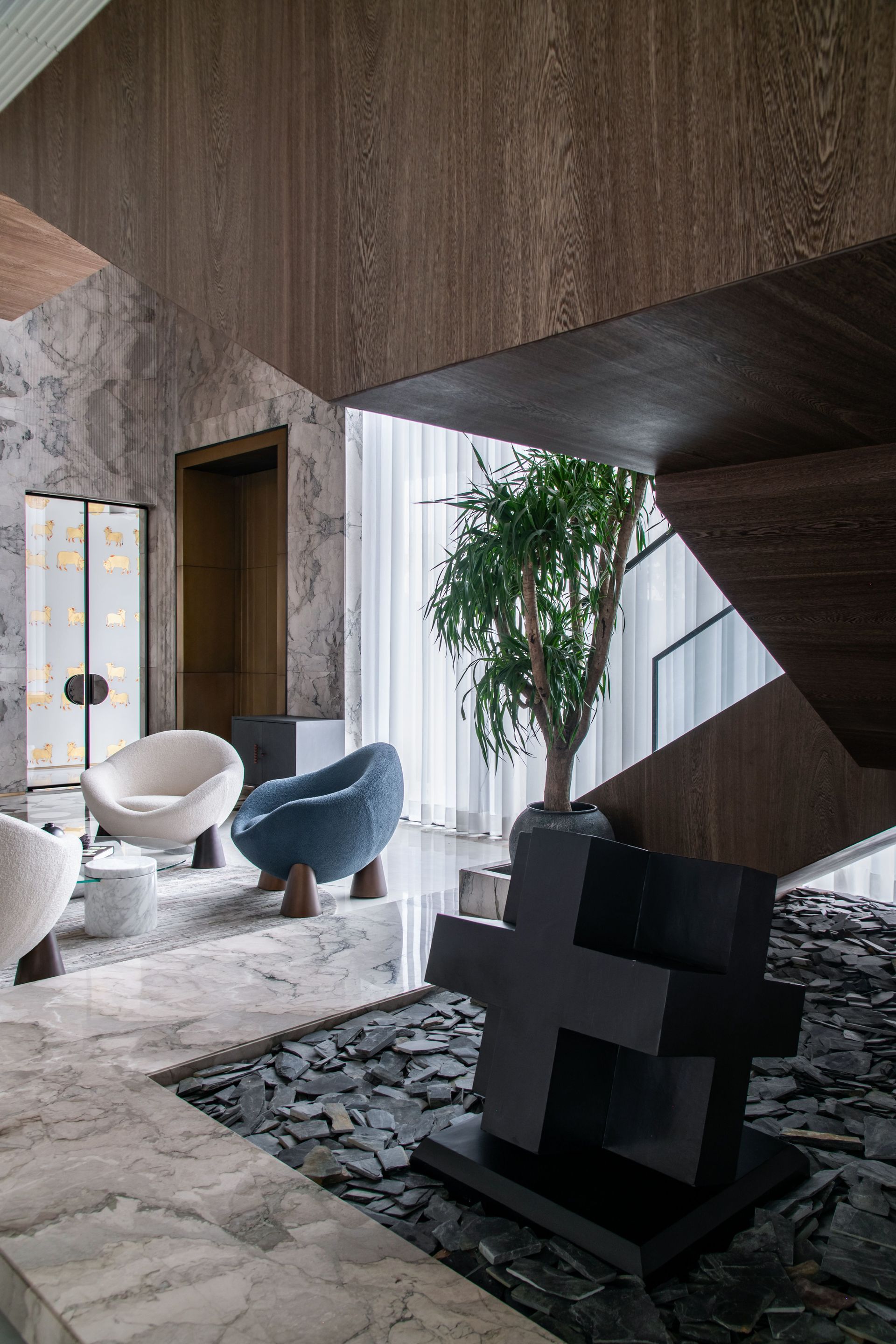 Modern lounge with sculptural chairs, wood-paneled walls, stone floor, and a potted tree by a large window