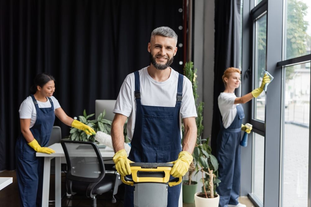 A man is holding a vacuum cleaner in an office while two women are cleaning windows.