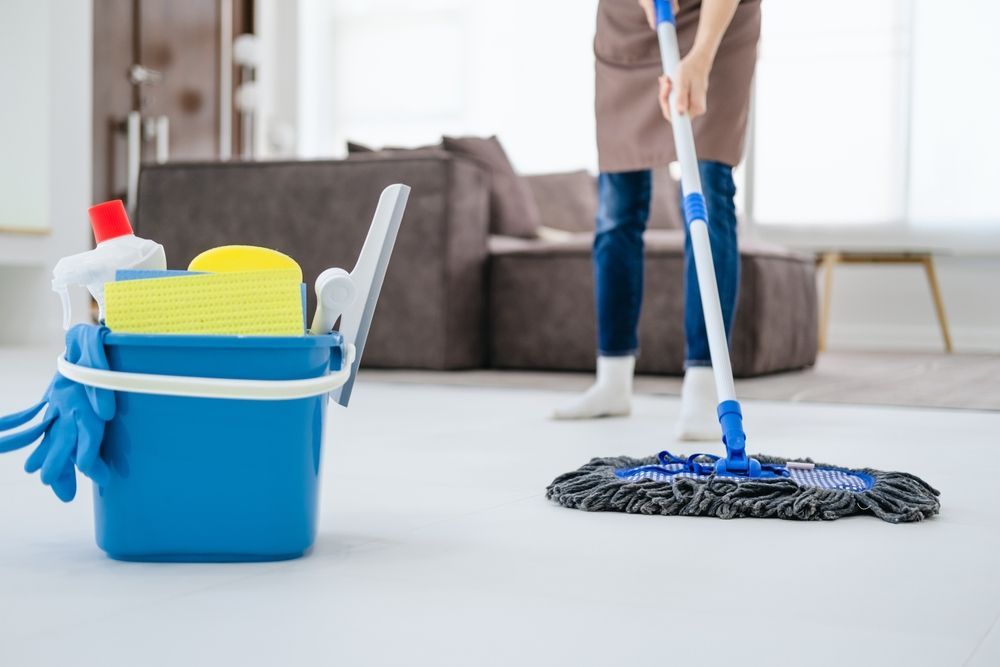 A woman is mopping the floor in a living room next to a bucket of cleaning supplies.