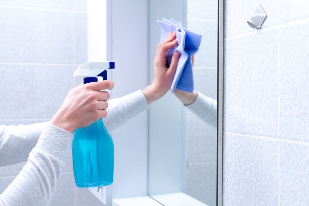 A person is cleaning a bathroom mirror with a spray bottle and a cloth.