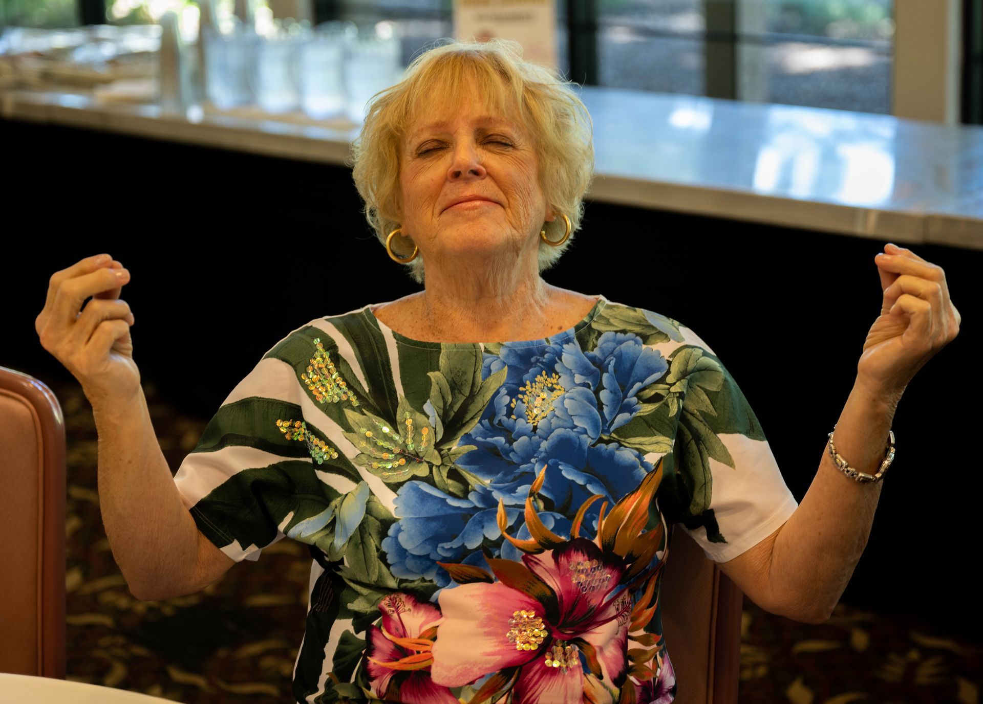 A woman in a floral dress is sitting at a table with her arms outstretched.