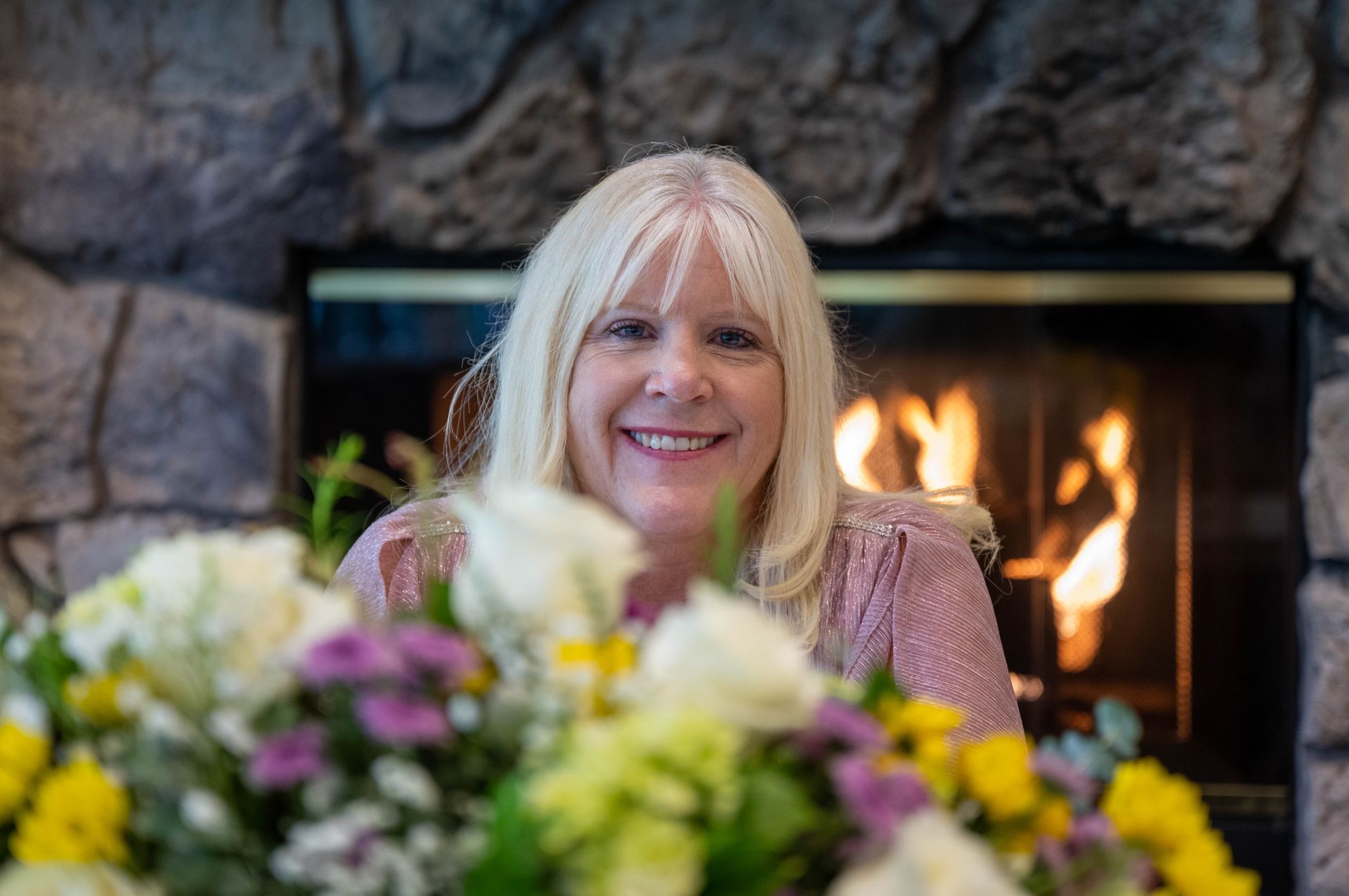 Kim Silverman is sitting in front of a fireplace with flowers in front of her.