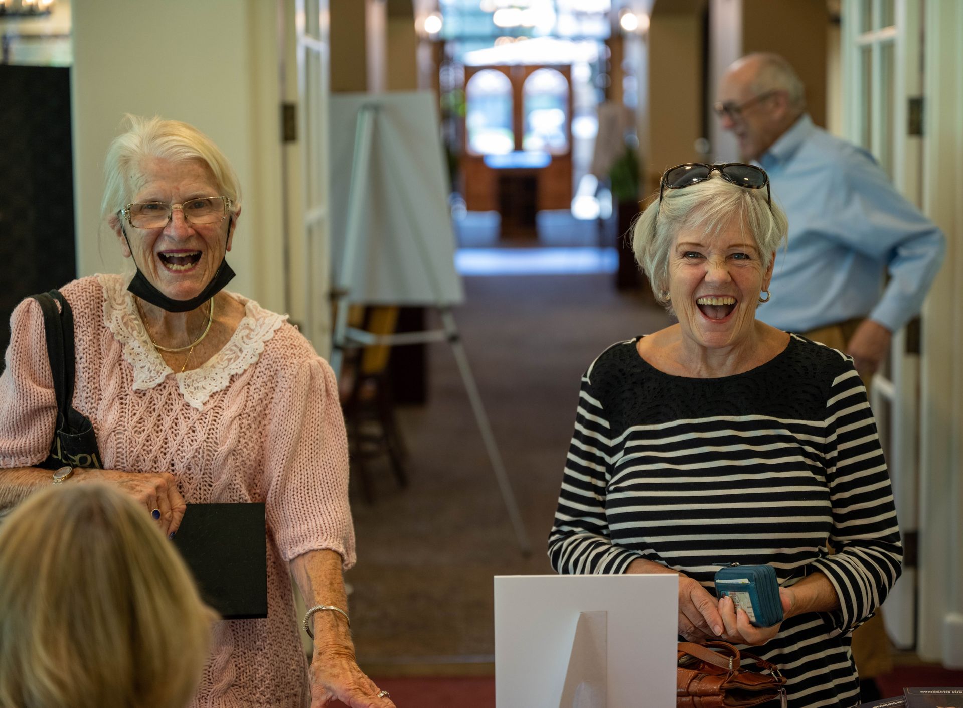 Two older women are standing next to each other in a hallway and smiling.