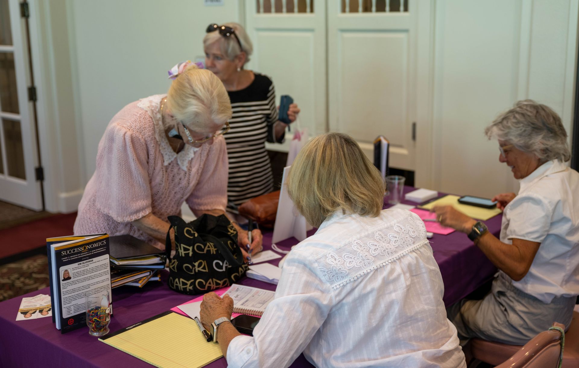 A group of women are sitting and standing at a table with a purple table cloth.