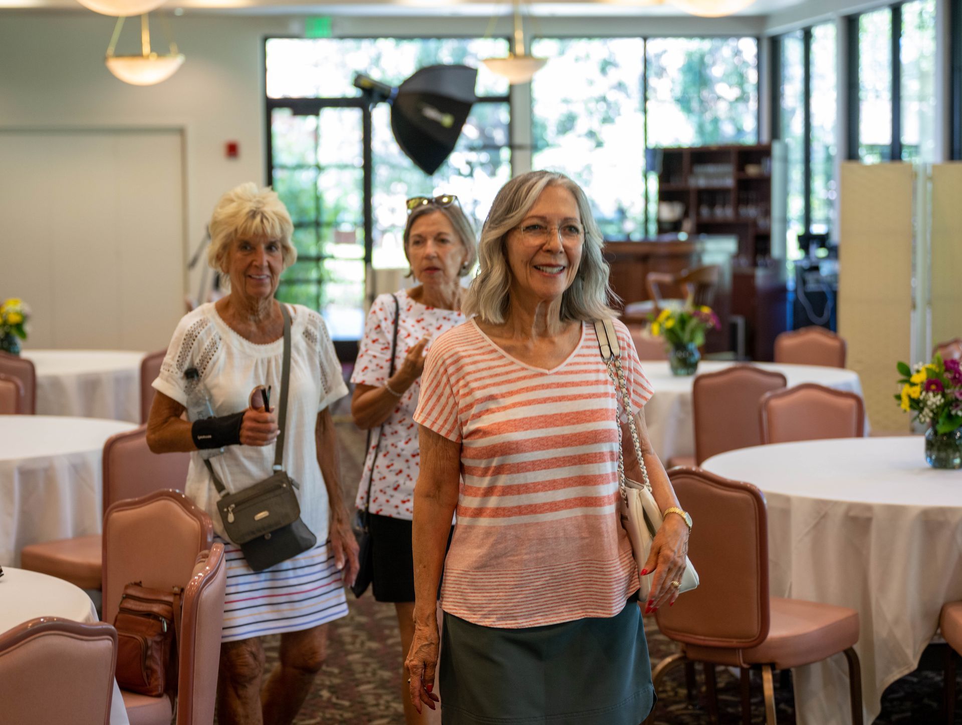 Three women are walking through a room with tables and chairs.