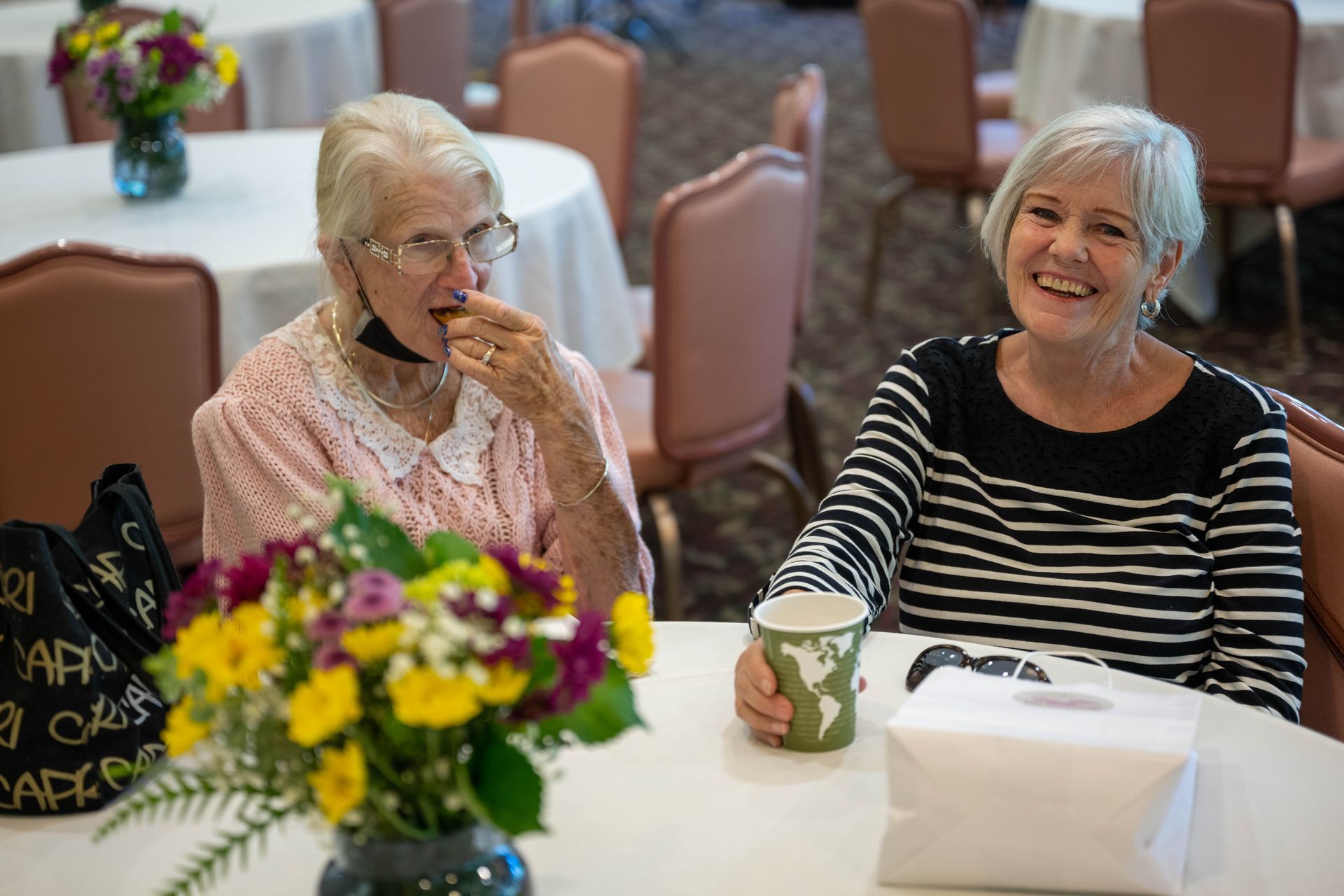 Two older women are sitting at a table with flowers and a cup of coffee.