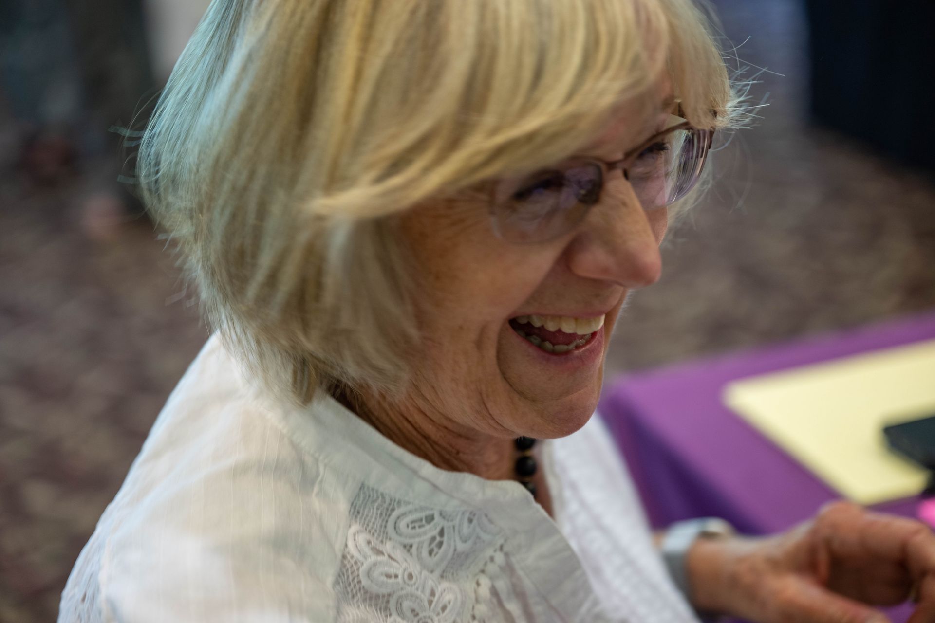 A woman wearing glasses and a white shirt is smiling while sitting at a table.