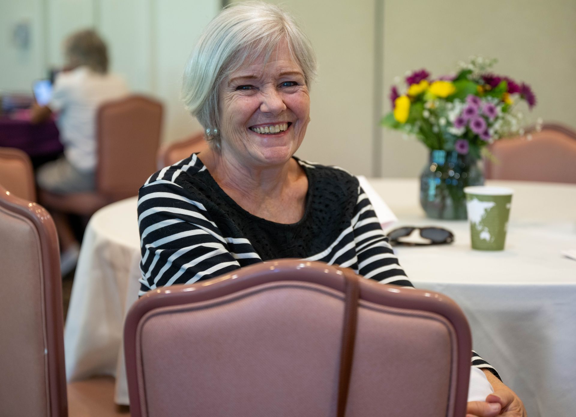 A woman is smiling while sitting at a table with flowers in the background.