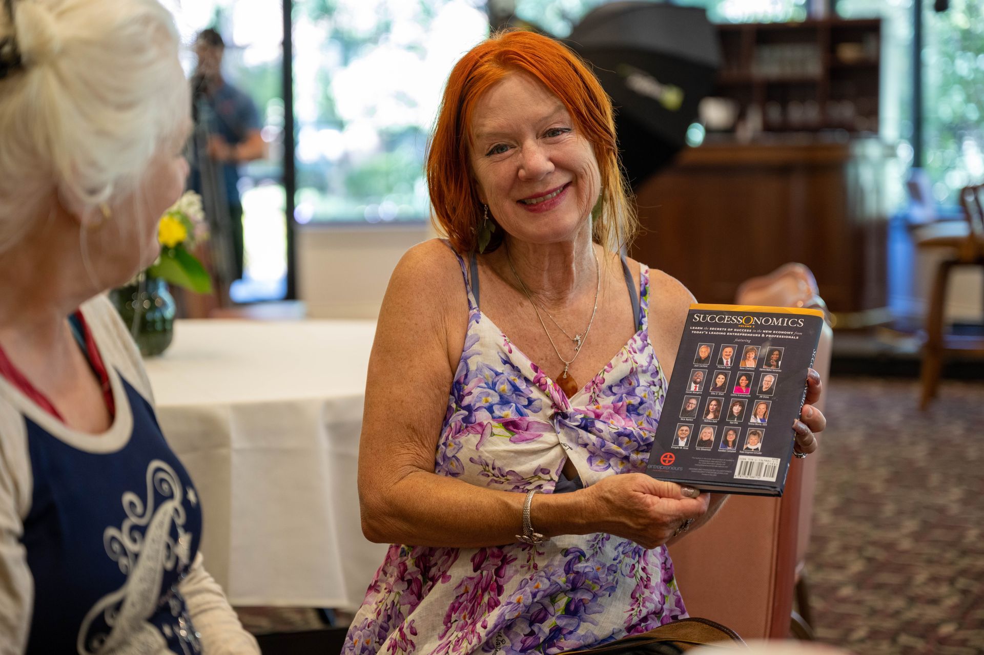 A woman is holding a book and smiling while sitting next to another woman.