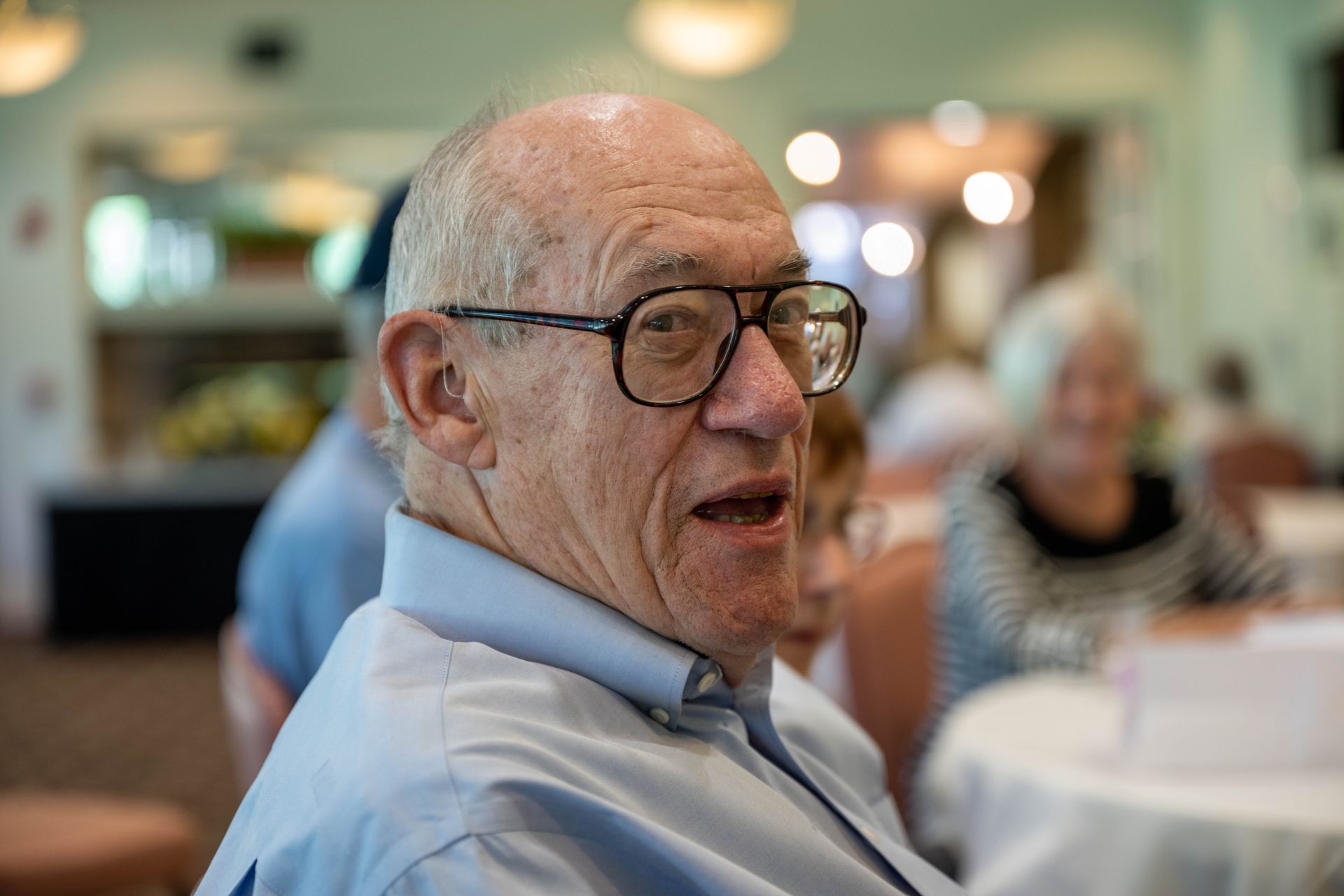 An elderly man wearing glasses is sitting at a table with his mouth open.