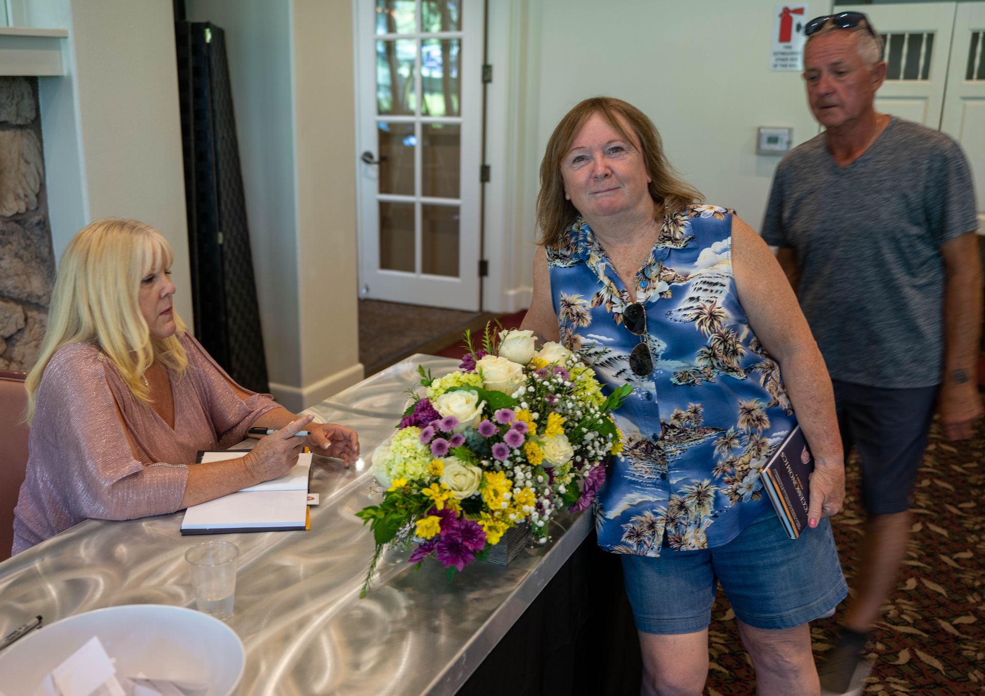 A woman is leaning on a table with a vase of flowers and a man is standing behind her.