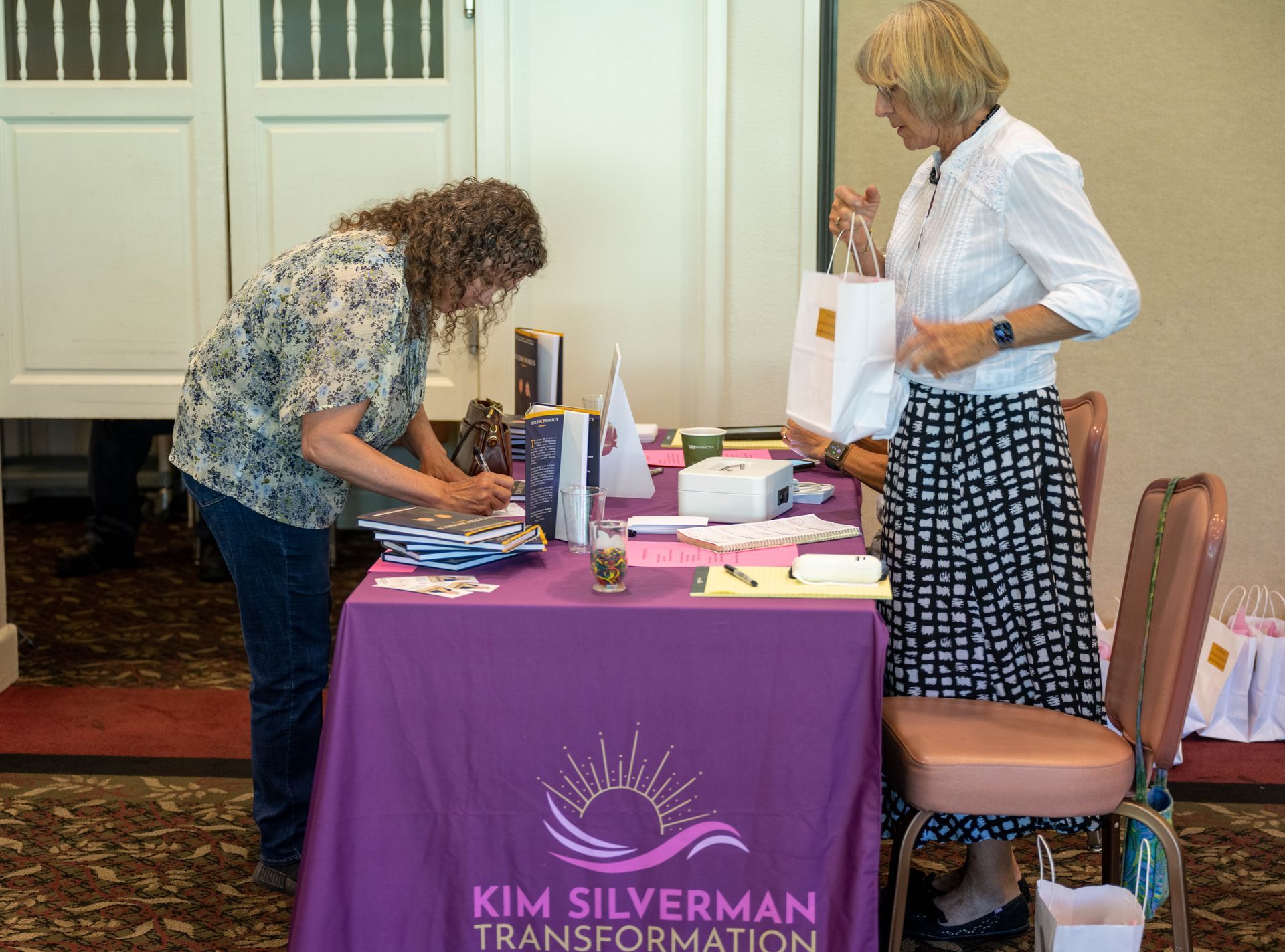 Two women are standing at a table with a purple table cloth.