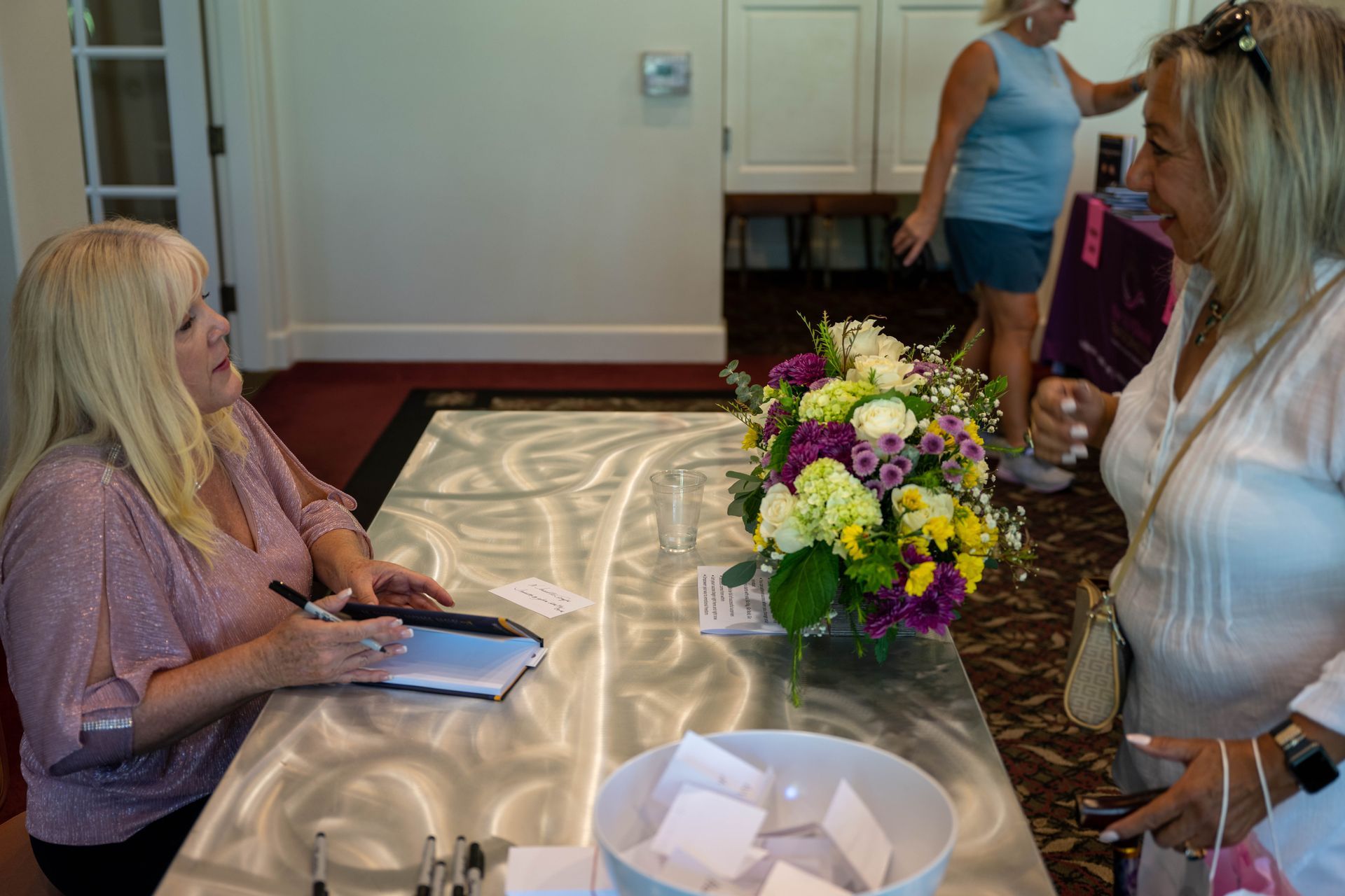 Two women are sitting at a table with a vase of flowers on it.