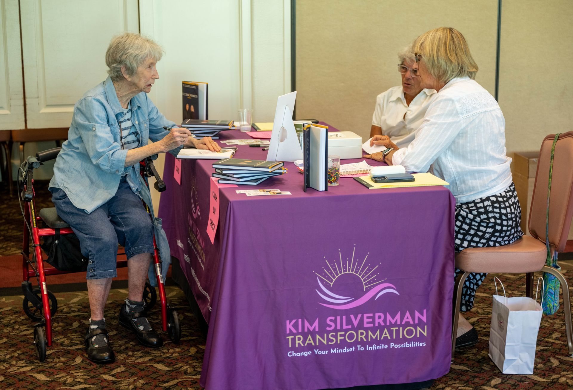 Three women are sitting at a table with a purple table cloth that says kim silverman transformation.