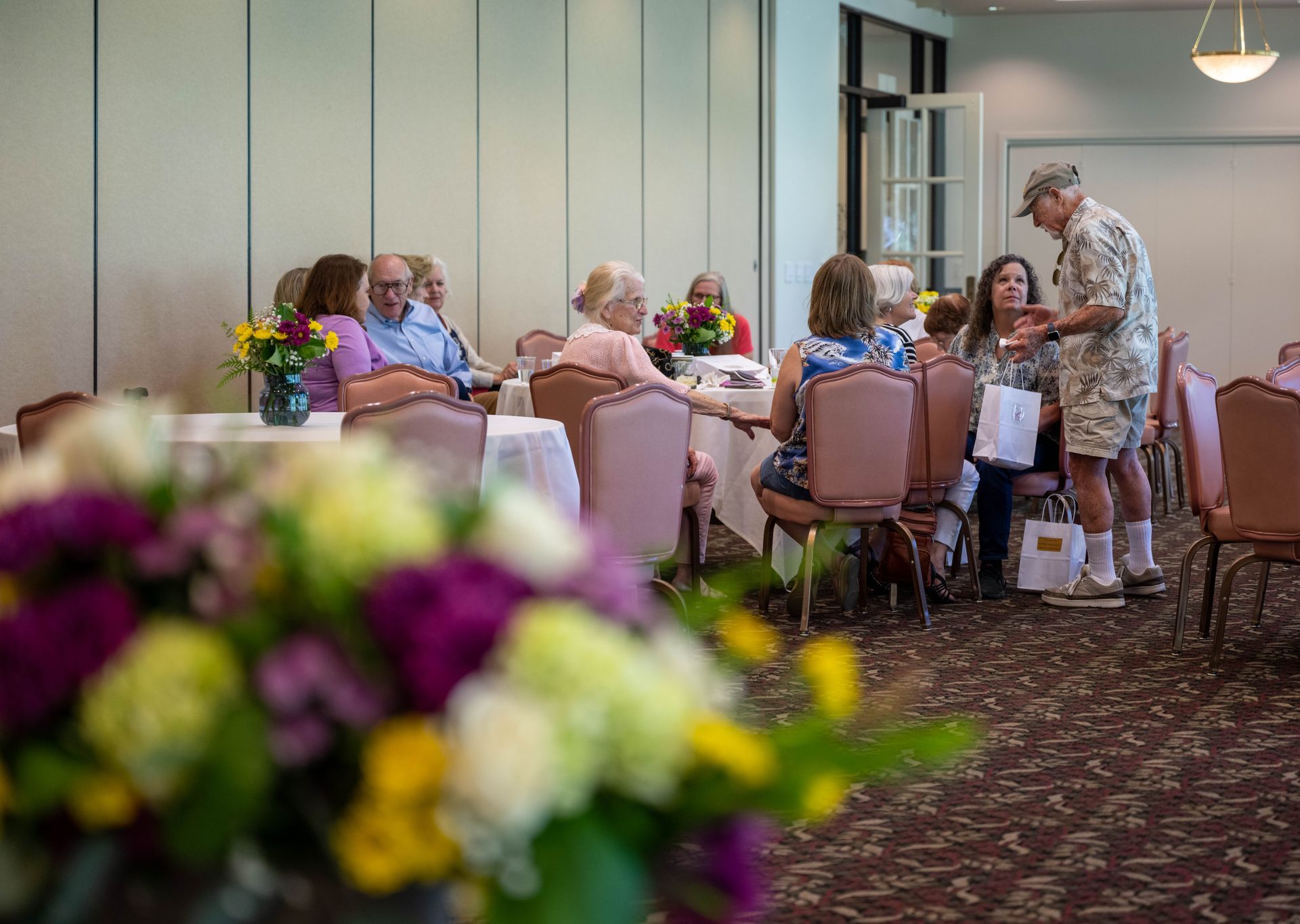 A group of people are sitting at tables in a room with flowers in the foreground.