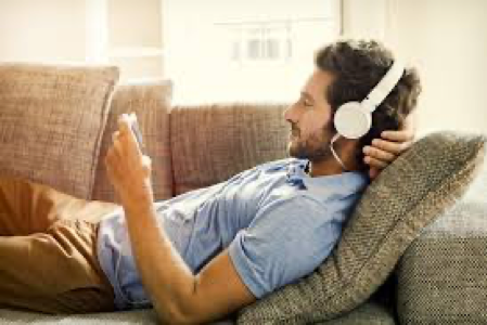 A man is laying on a couch wearing headphones and using a tablet.