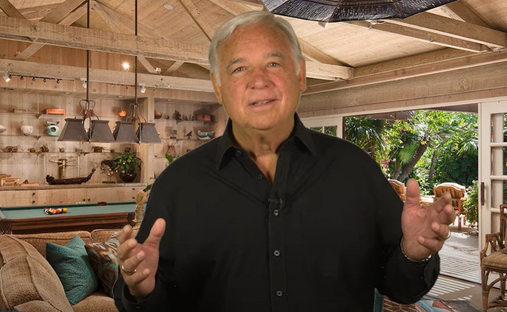 Jack Canfield in a black shirt is giving a thumbs up in a living room.
