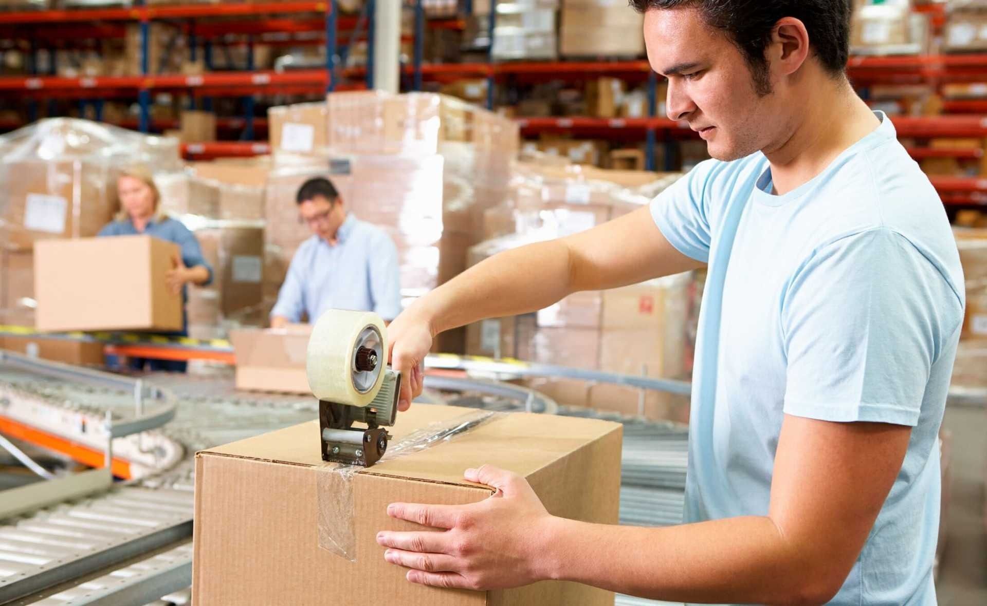 Un hombre está pegando con cinta adhesiva una caja de cartón en un almacén.