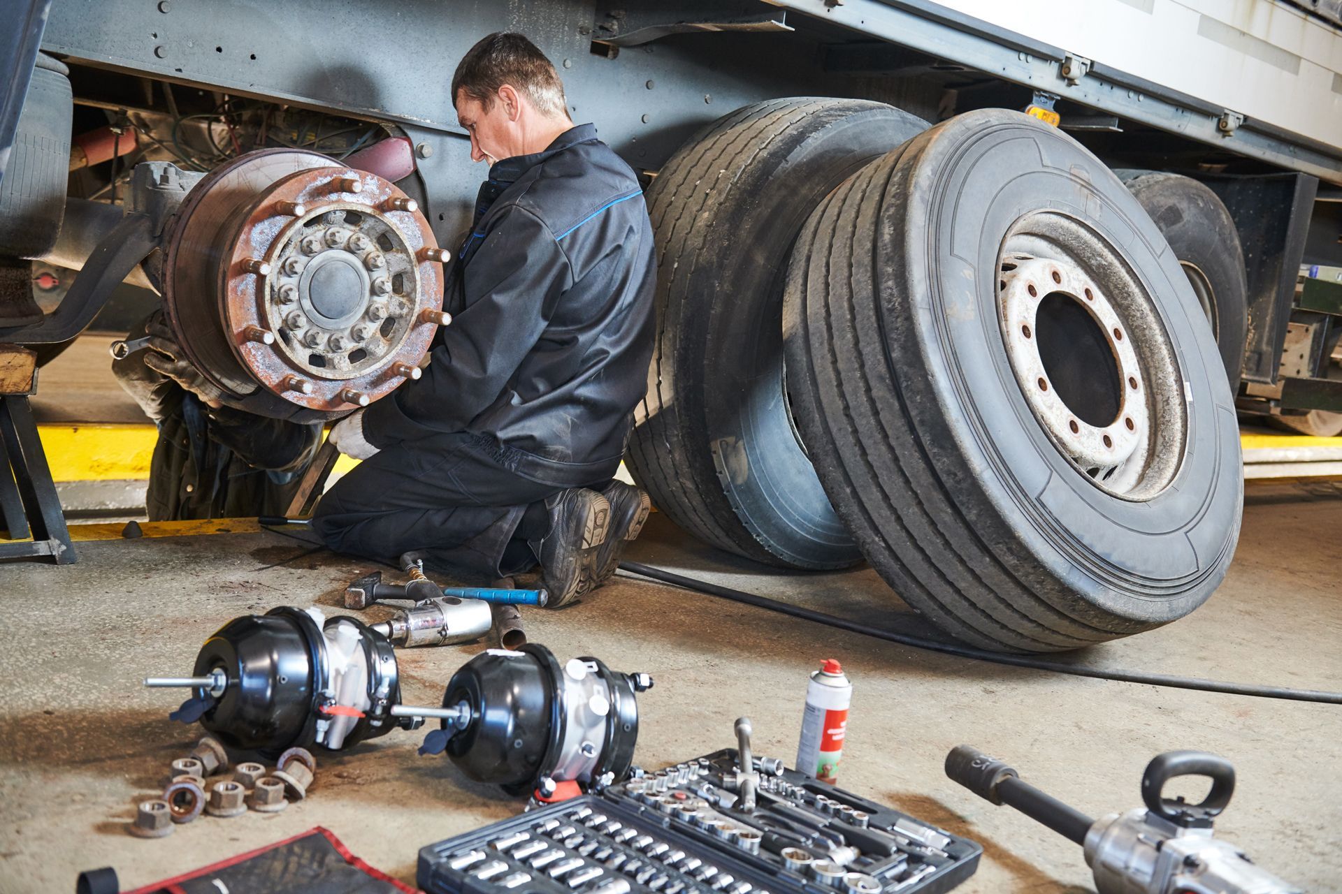 A mechanic works on a truck's wheel assembly in a repair shop with tools and spare brake parts on the floor nearby.