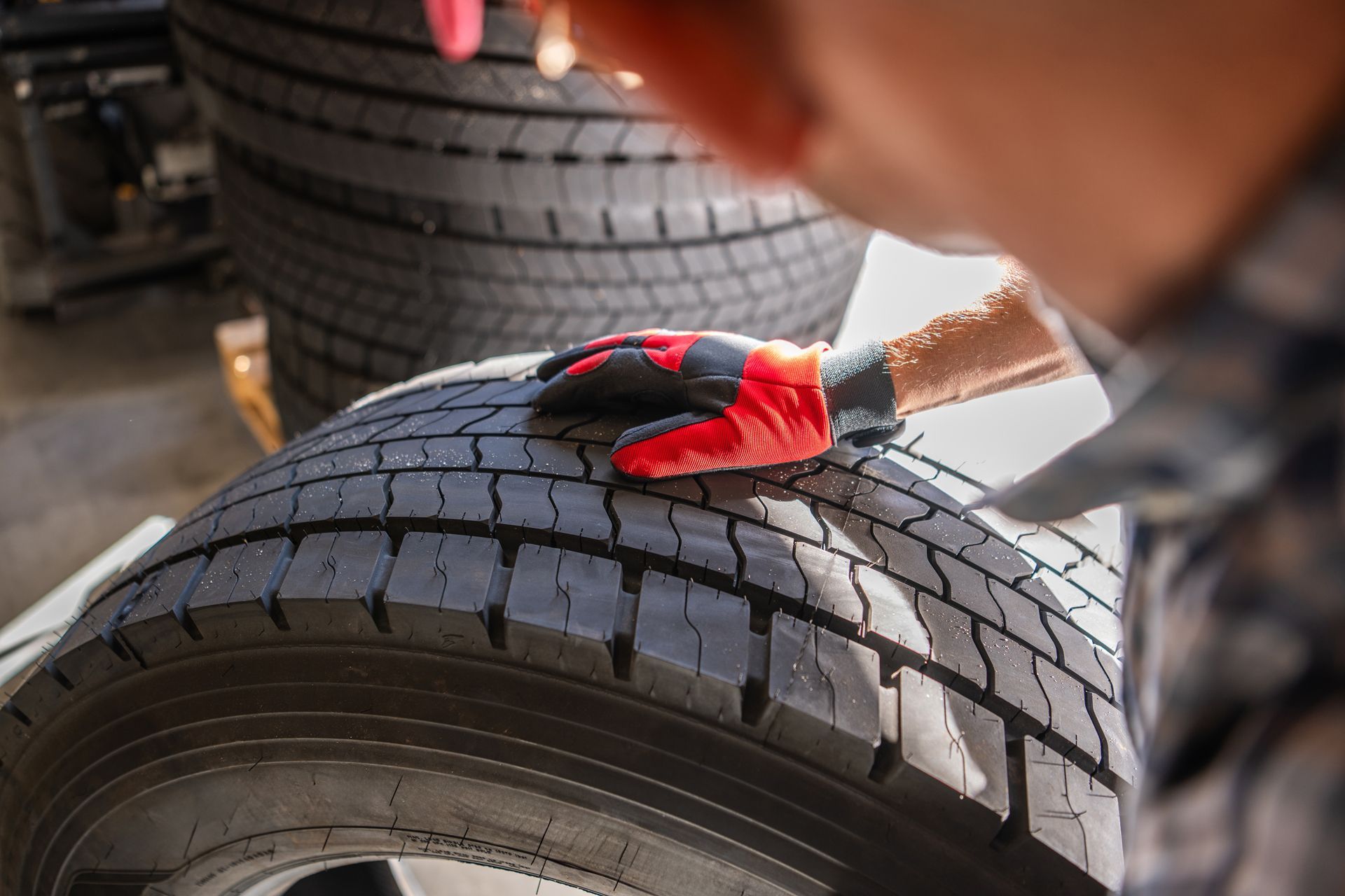 A person wearing a red and black work glove examines the deep tread pattern on a large black tire in a shop.