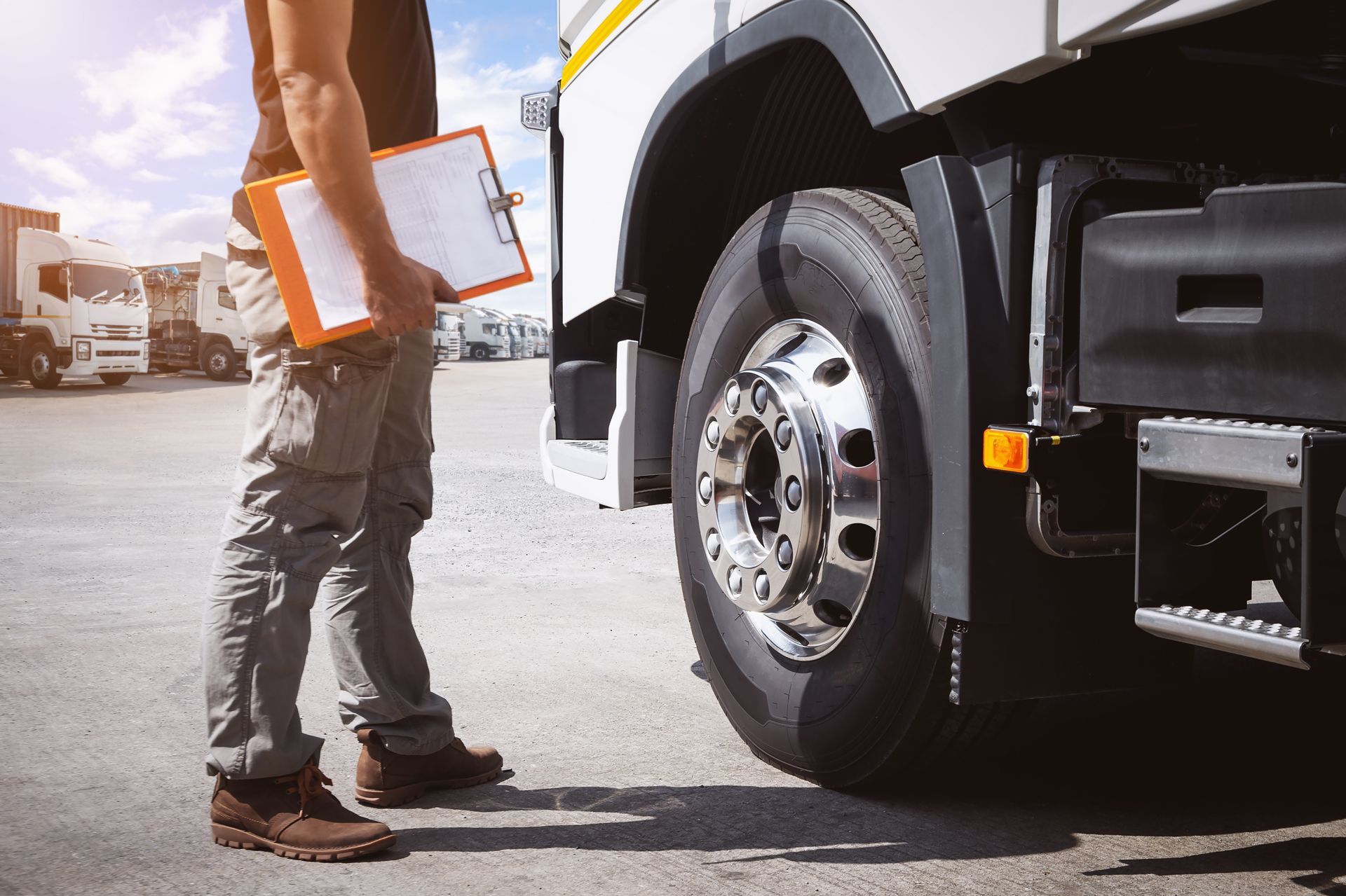 A person holding a clipboard stands next to the large front wheel of a semi-truck in a parking lot.