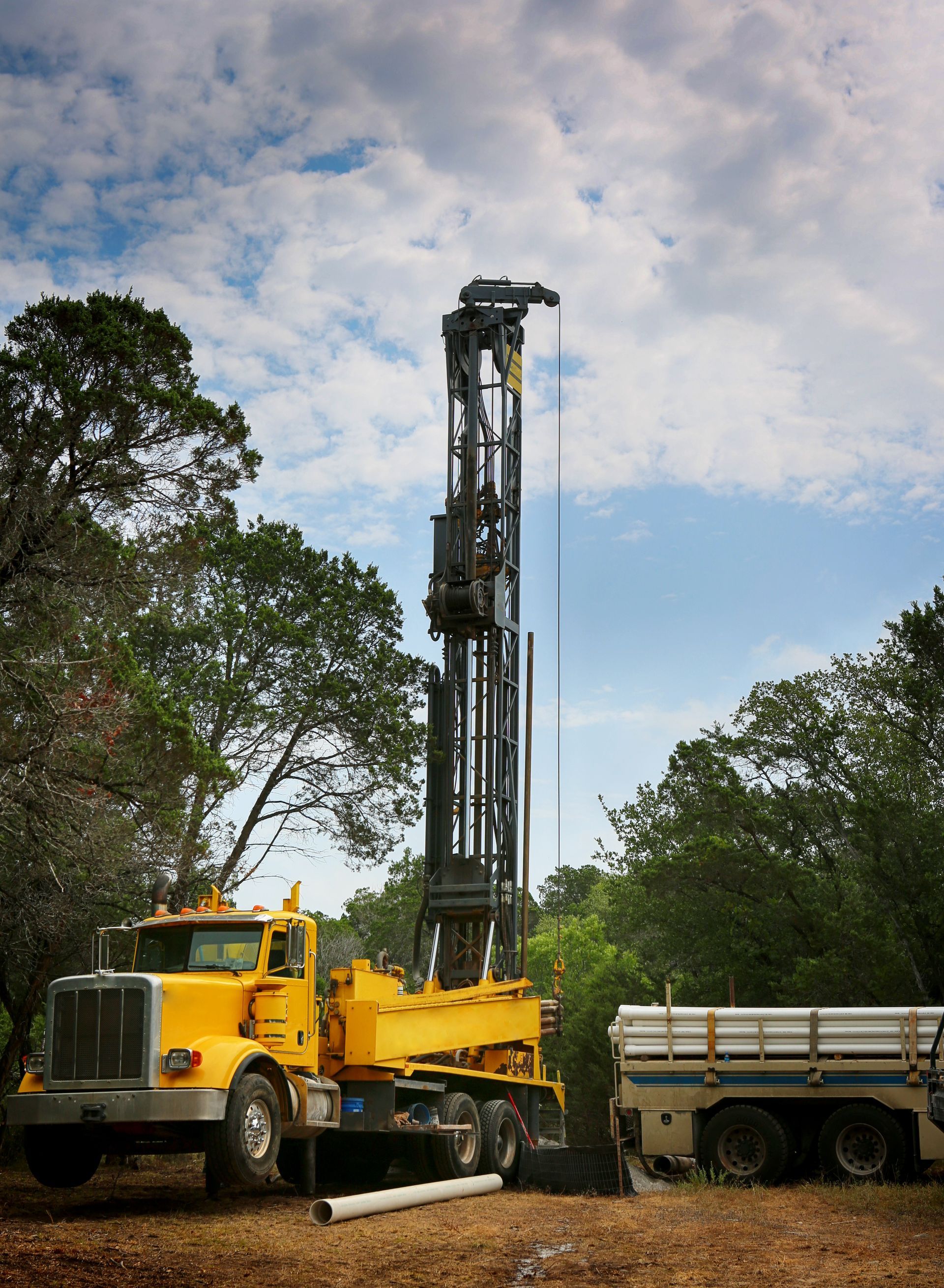 Yellow drilling rig truck beside a white trailer in a wooded area under a cloudy sky