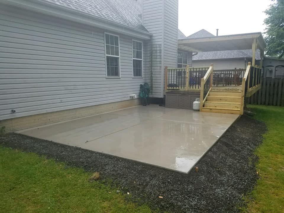 Backyard with a house, patio, and overgrown garden on a slight slope, with a brown fence on the left.