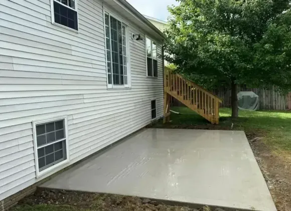 Gray concrete patio next to a house with white siding and a wooden staircase leading up to a deck.