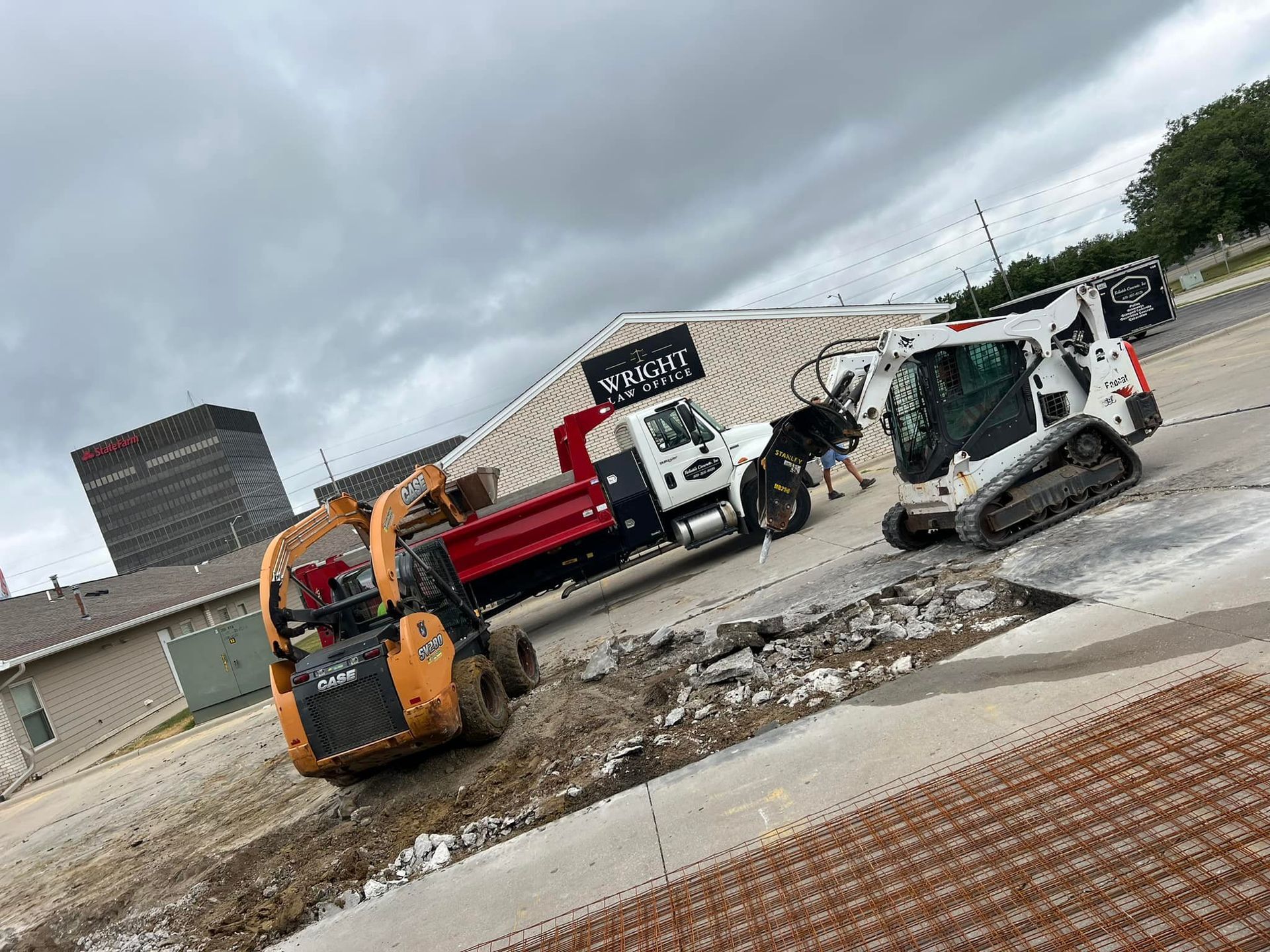 Yellow excavator on a pile of gravel in a wooded area with trees and a cloudy sky.