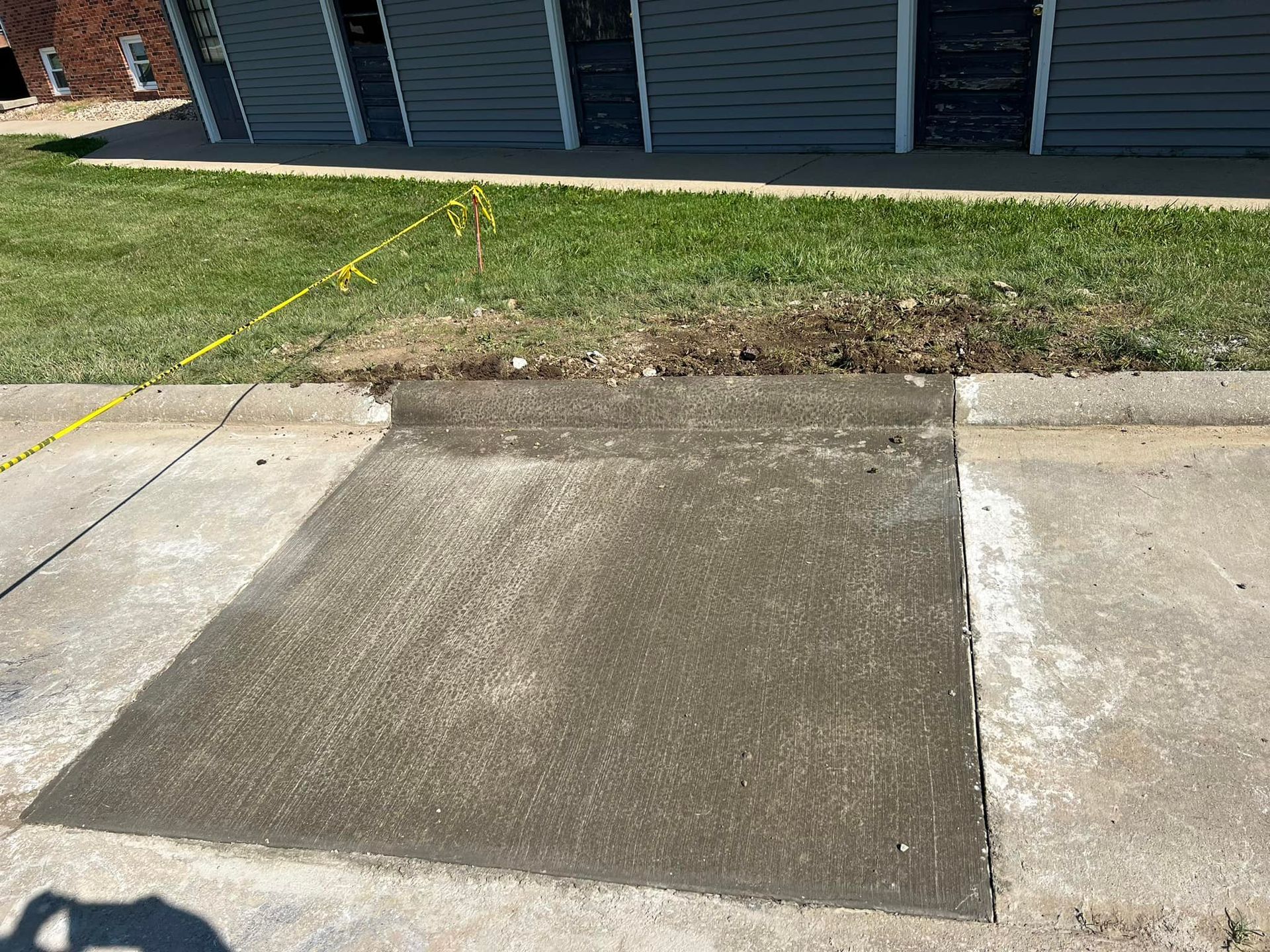 Newly poured concrete sidewalk patch in front of a building with grass and yellow caution tape.
