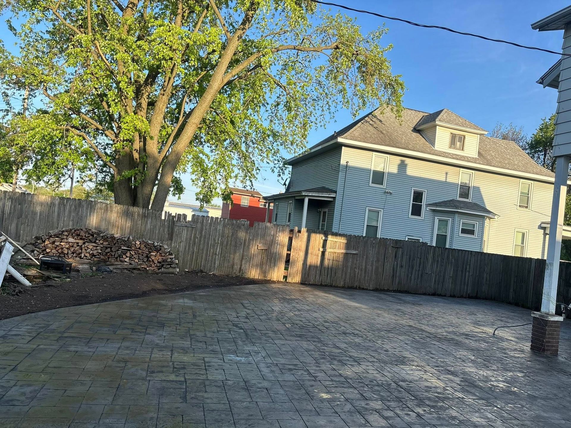 A backyard with a stamped concrete patio, wooden fence, house in the background, and tree.