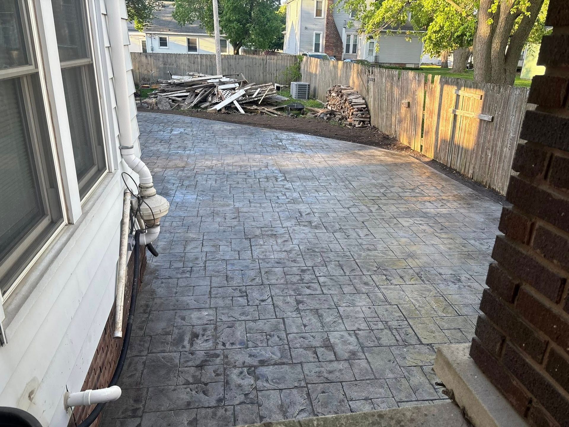 Concrete patio with textured, brick-like pattern next to a house with a wooden fence in the background.