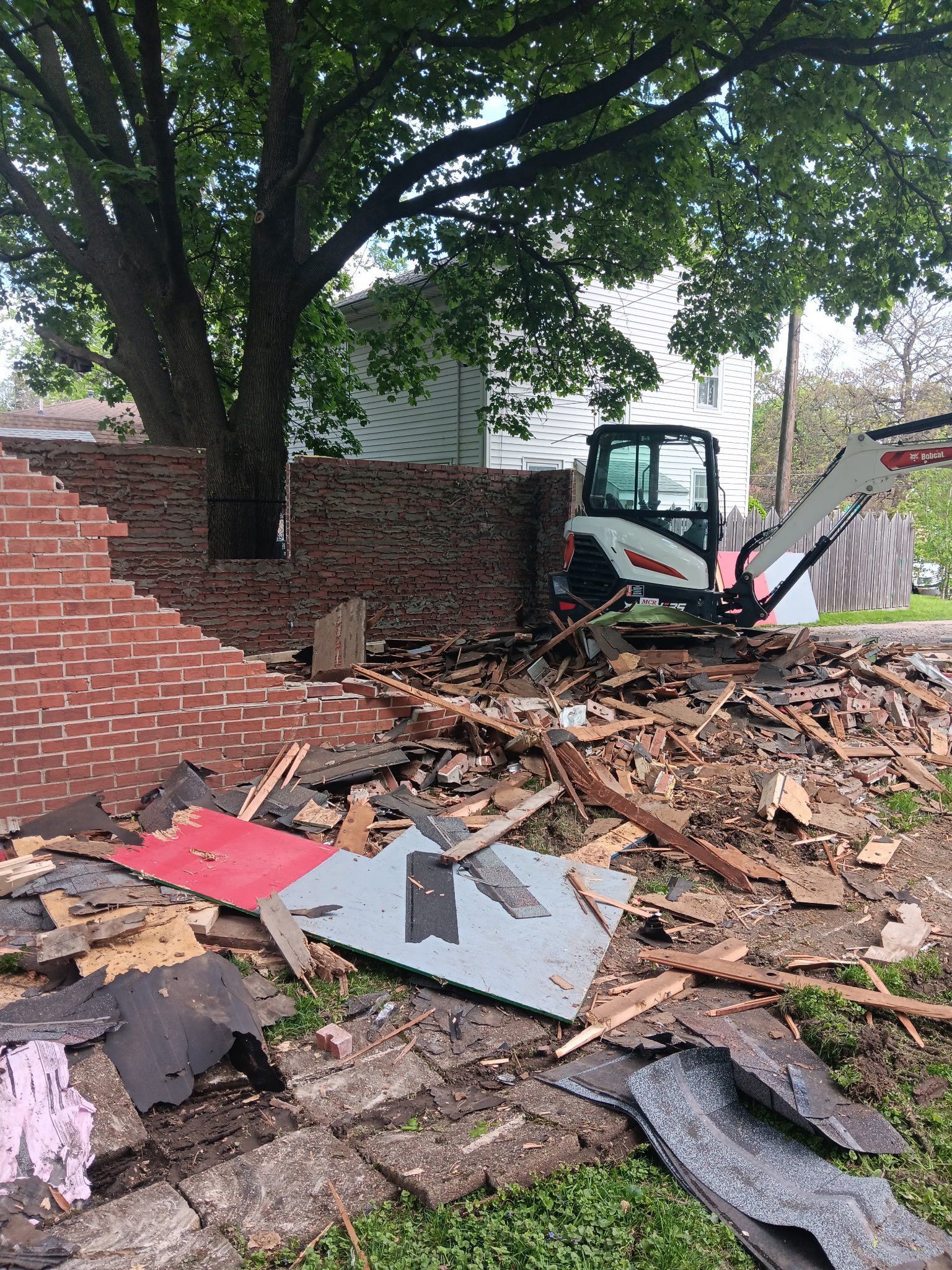 Bobcat machine demolishing brick wall by tree, surrounded by debris.