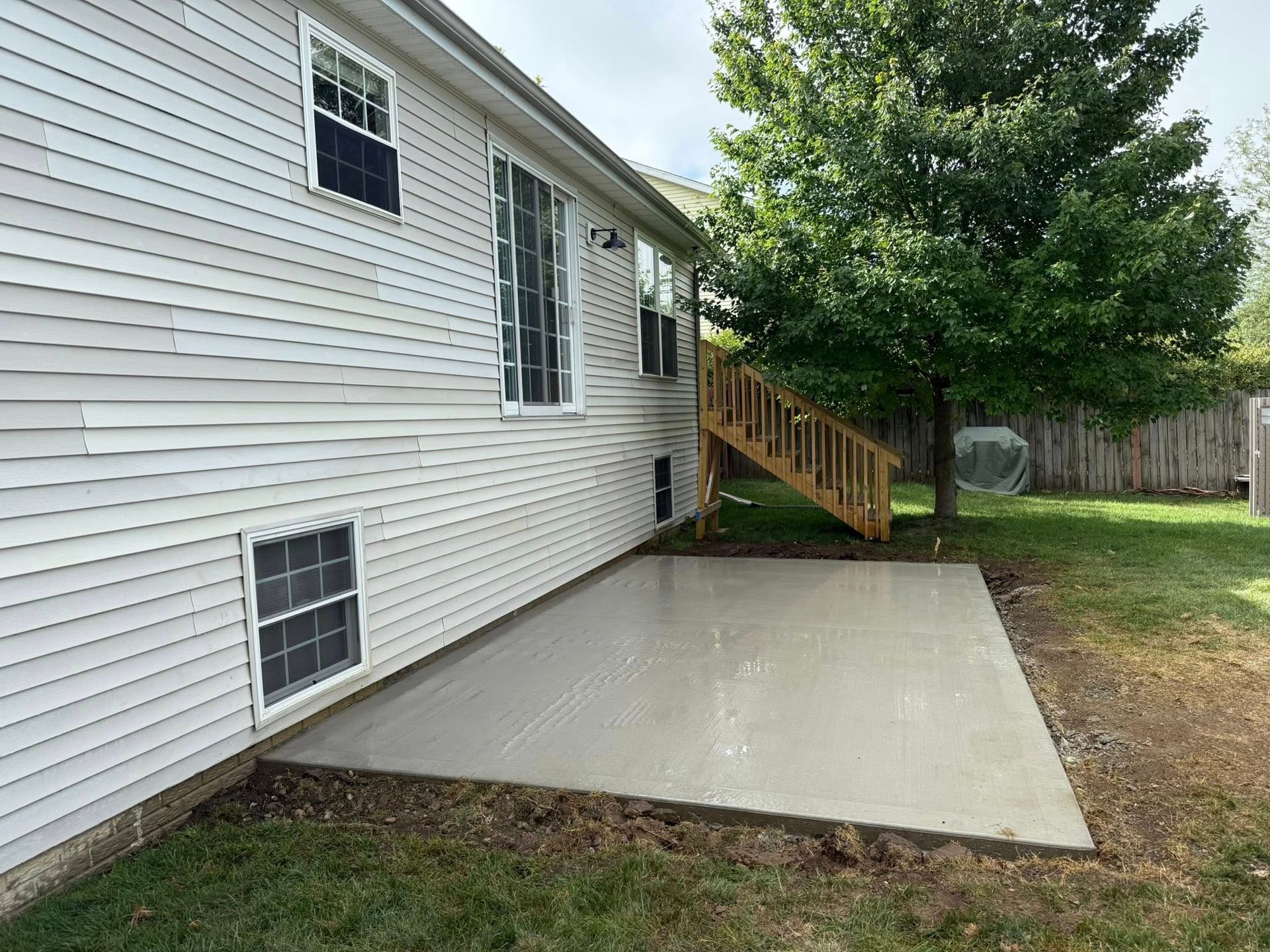 Side view of a house with a concrete patio. Stairs lead to a wooden deck. A tree and grass surround the house.
