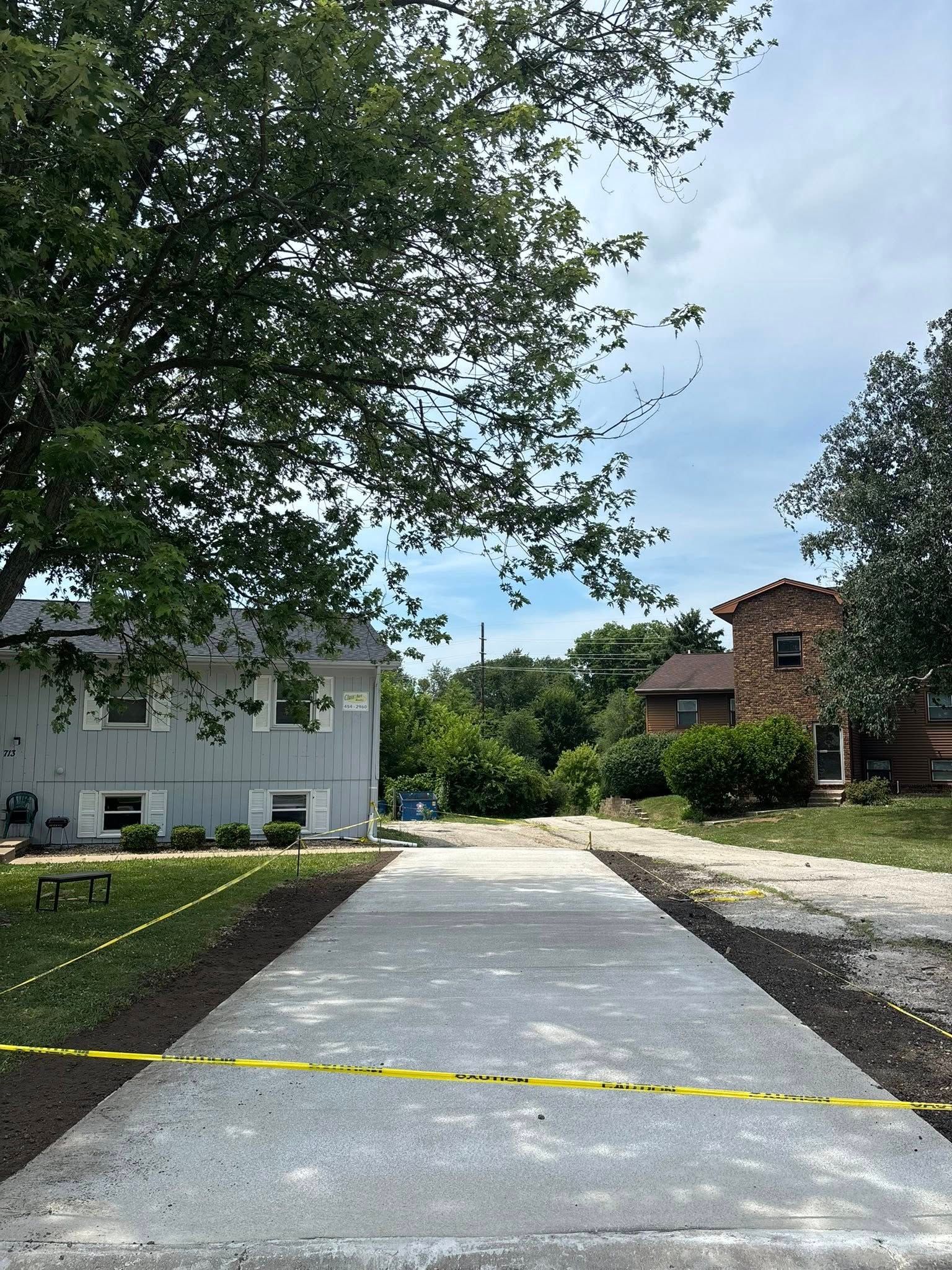 Newly paved concrete driveway leading towards houses under a cloudy sky, with caution tape and trees.