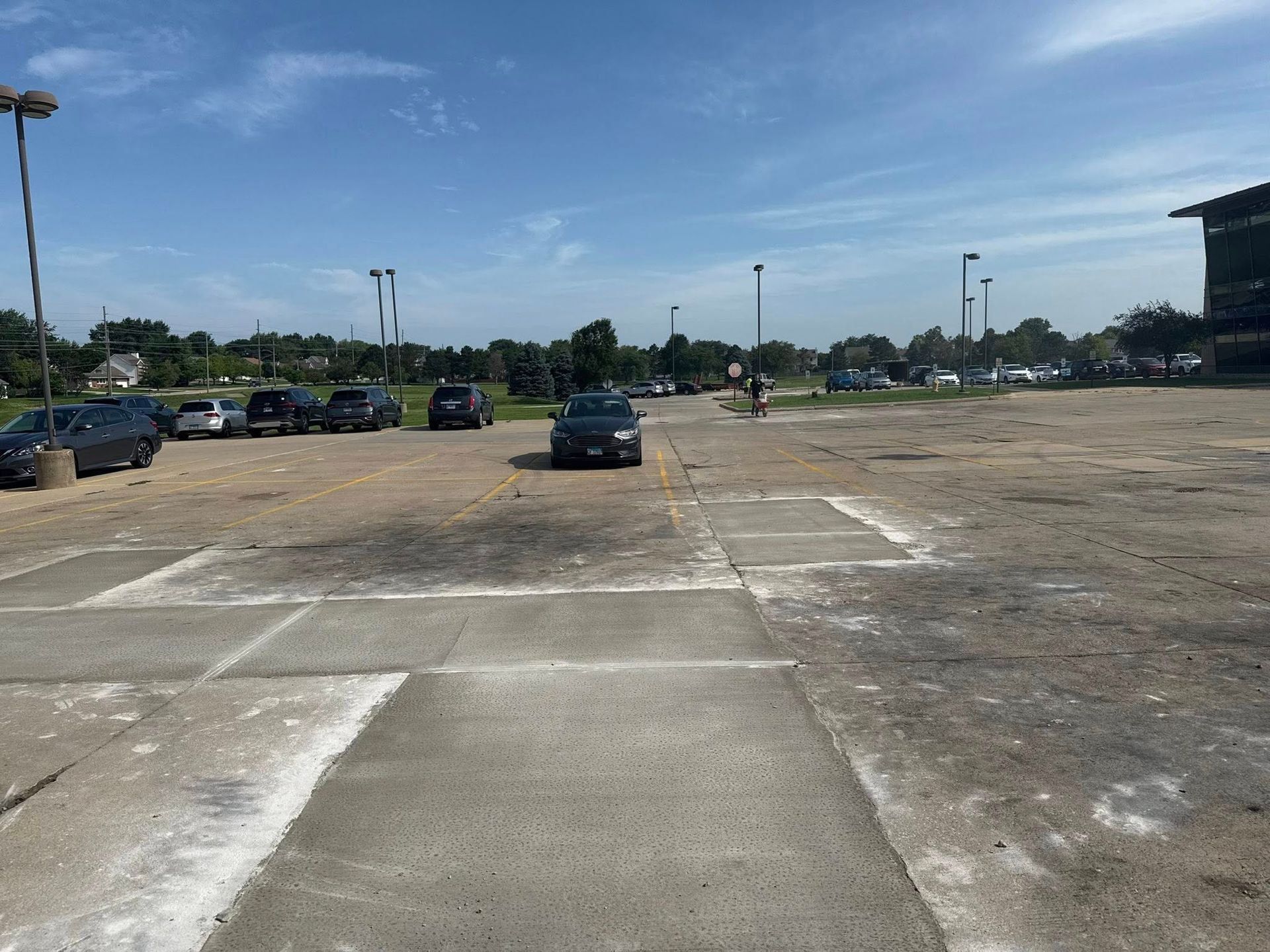 Parking lot with scattered cars, mostly empty, under a blue sky with some trees and a building in the background.