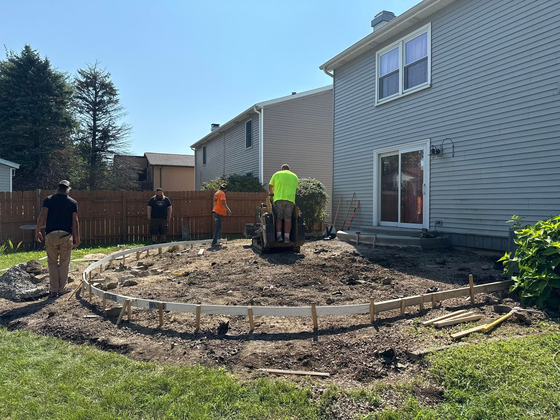 Construction workers building a curved concrete border in a backyard. A small excavator is in use.