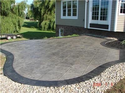 Gray brick-patterned concrete patio next to a white house with a pile of wood in the background.