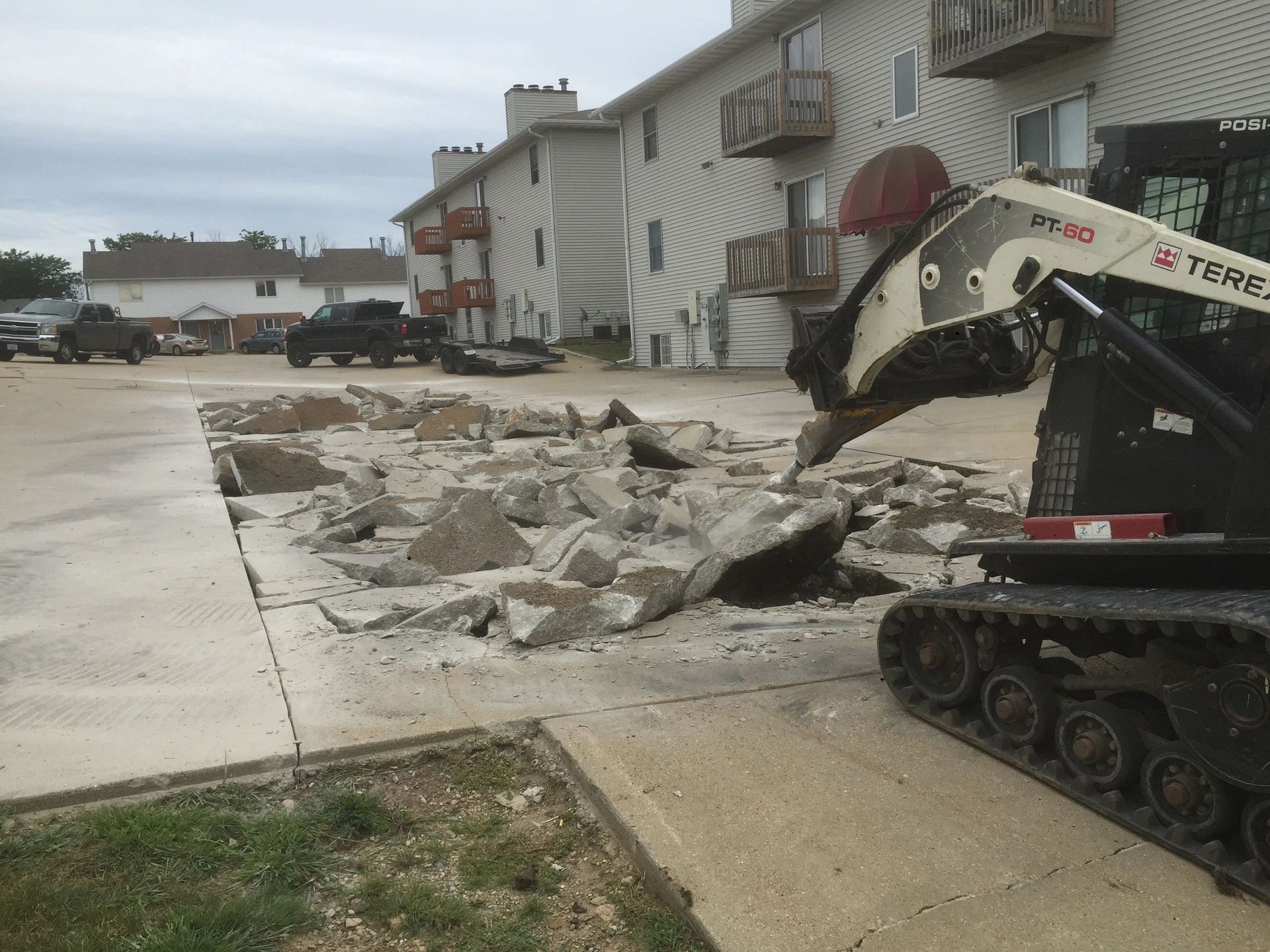 Construction vehicle demolishing concrete in front of an apartment building on a cloudy day.