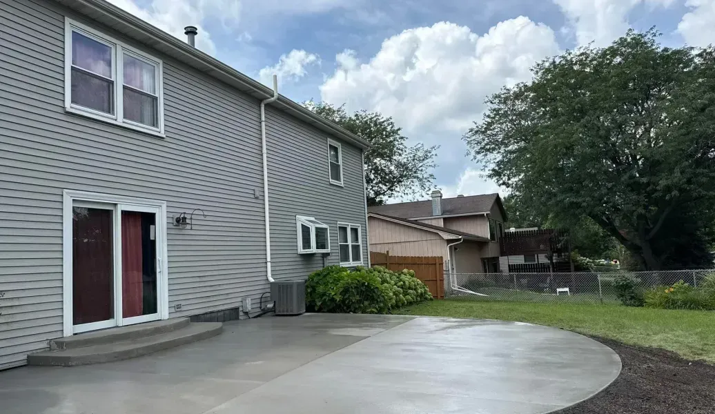 Gray two-story house with a circular concrete patio in a backyard setting, cloudy sky.