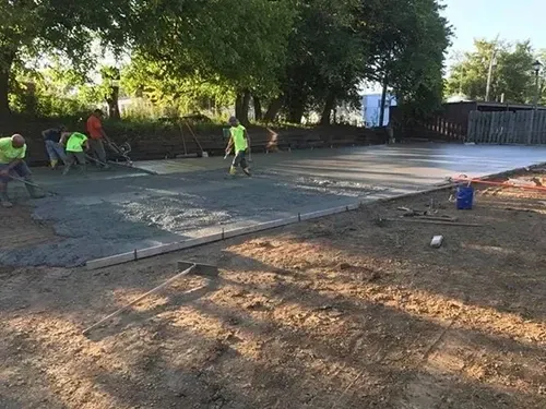 Workers pouring and smoothing concrete on a construction site. Green and orange vests are visible.