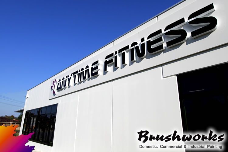 An Anytime Fitness building with a white exterior, black windows, and a blue sky. The logo is on the front and a logo for Brushworks at the bottom — Brushworks Painting In North Albury, NSW.