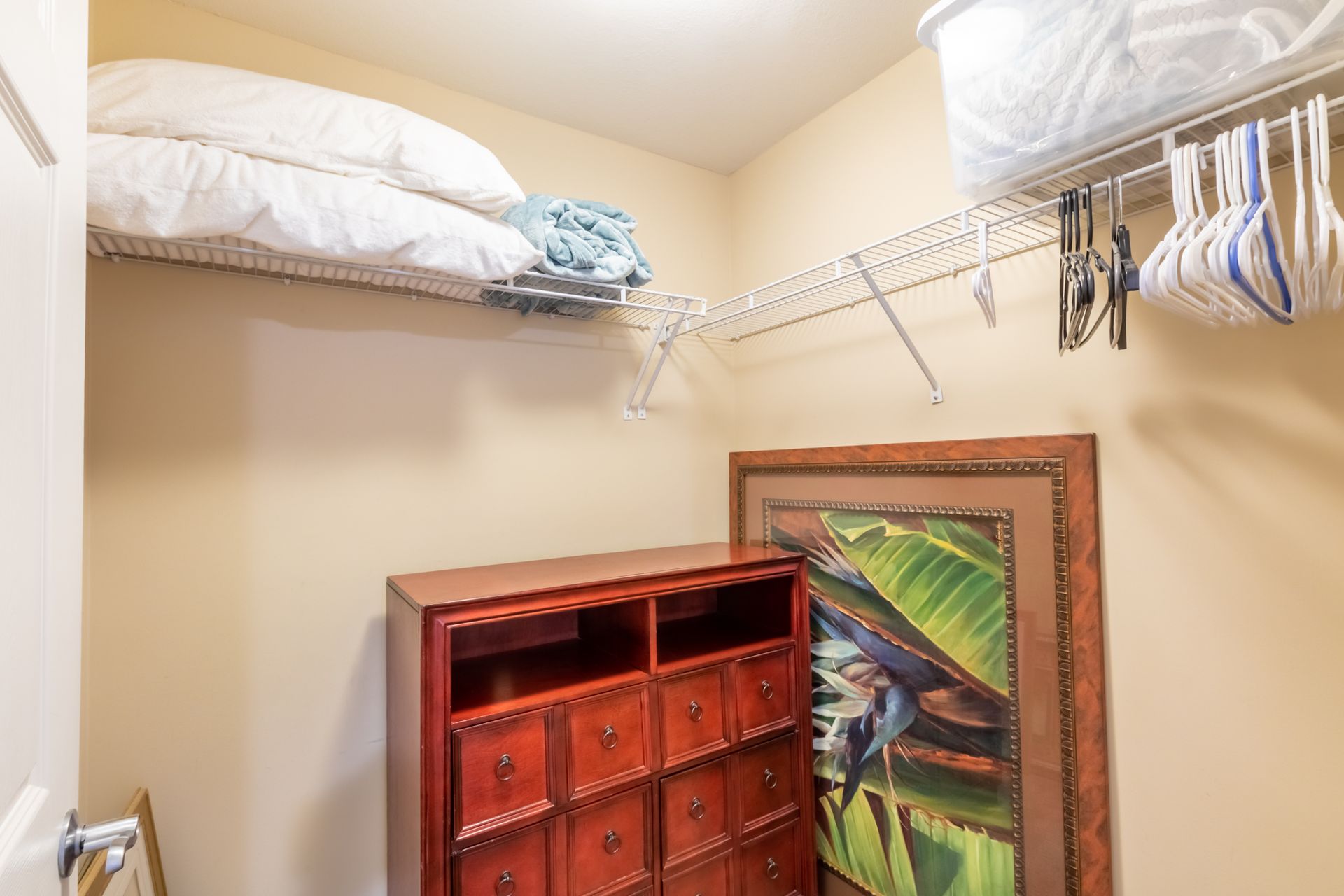 A small storage closet with a dresser, large framed artwork, and a shelf holding blankets and hangers.