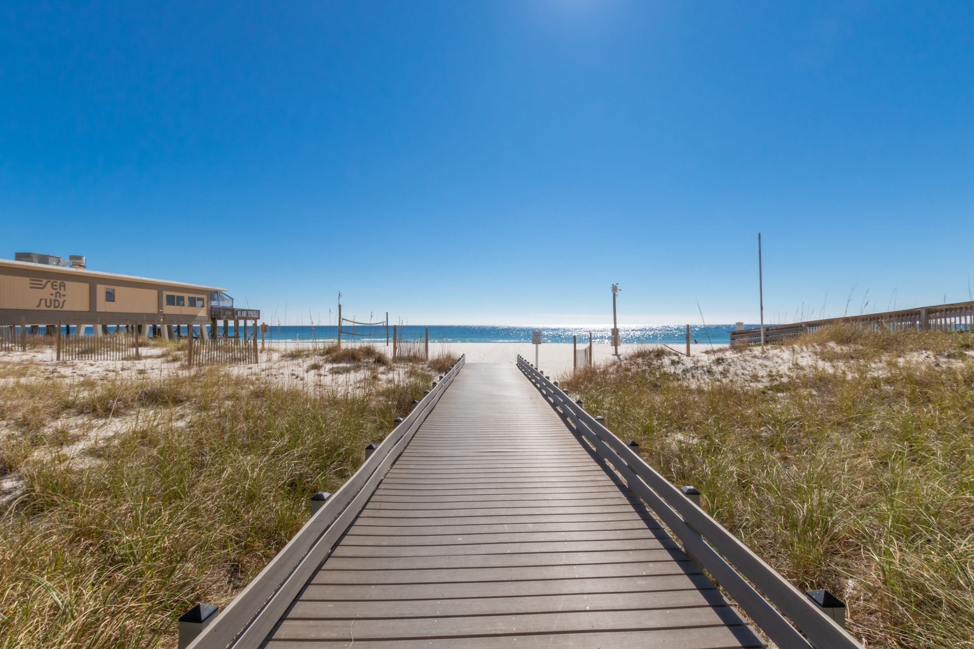 Wooden boardwalk to white sandy beach under a bright blue sky.