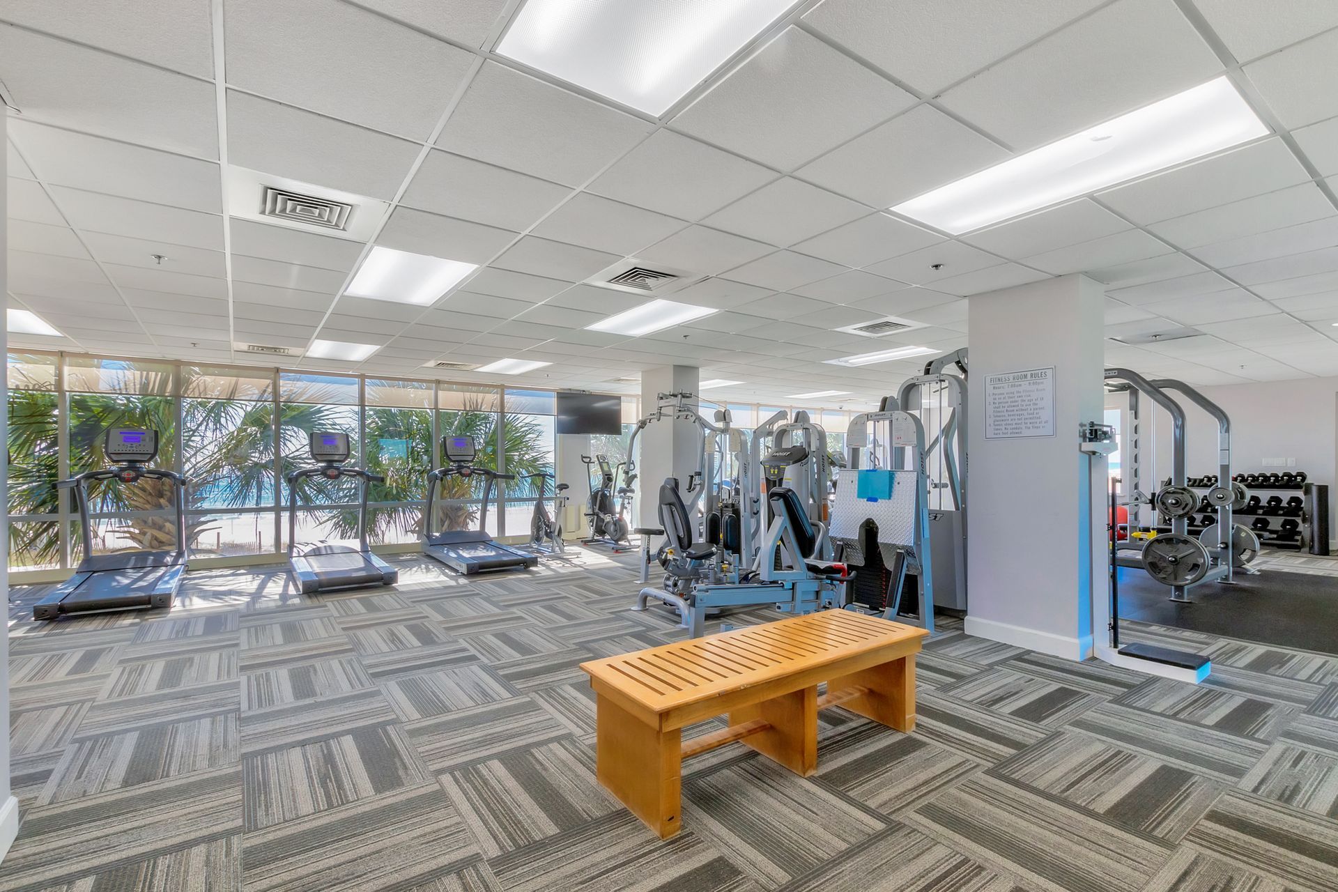 Gym interior with various exercise machines, windows, and a wooden bench.