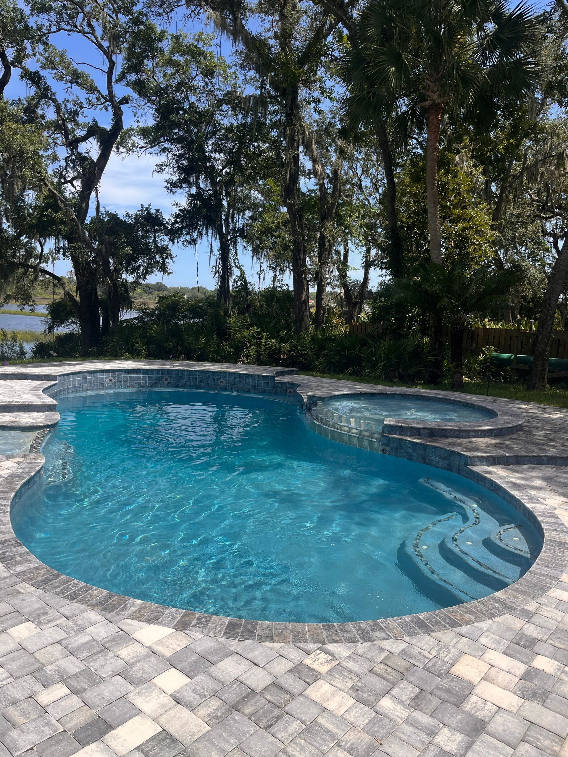 Swimming pool with blue water, built-in hot tub, surrounded by gray pavers, trees in background.