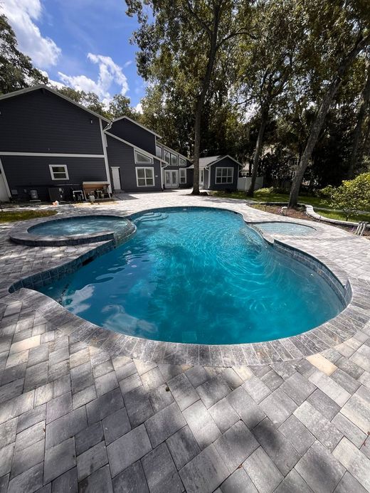 A backyard with a pool, spa, and brick patio, next to a dark gray house.