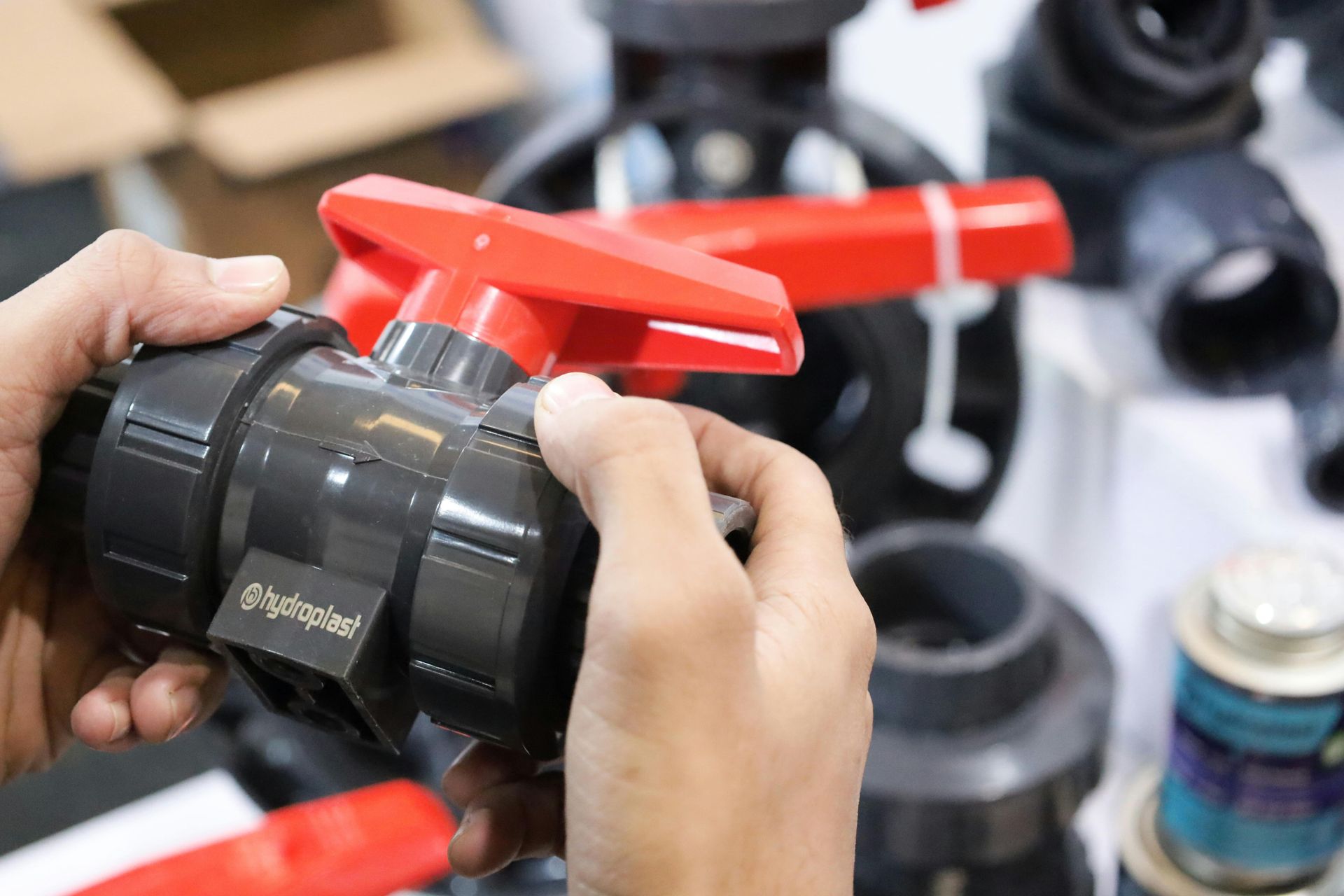 Hands holding a dark gray plastic valve with a red handle, other plumbing parts in the background.