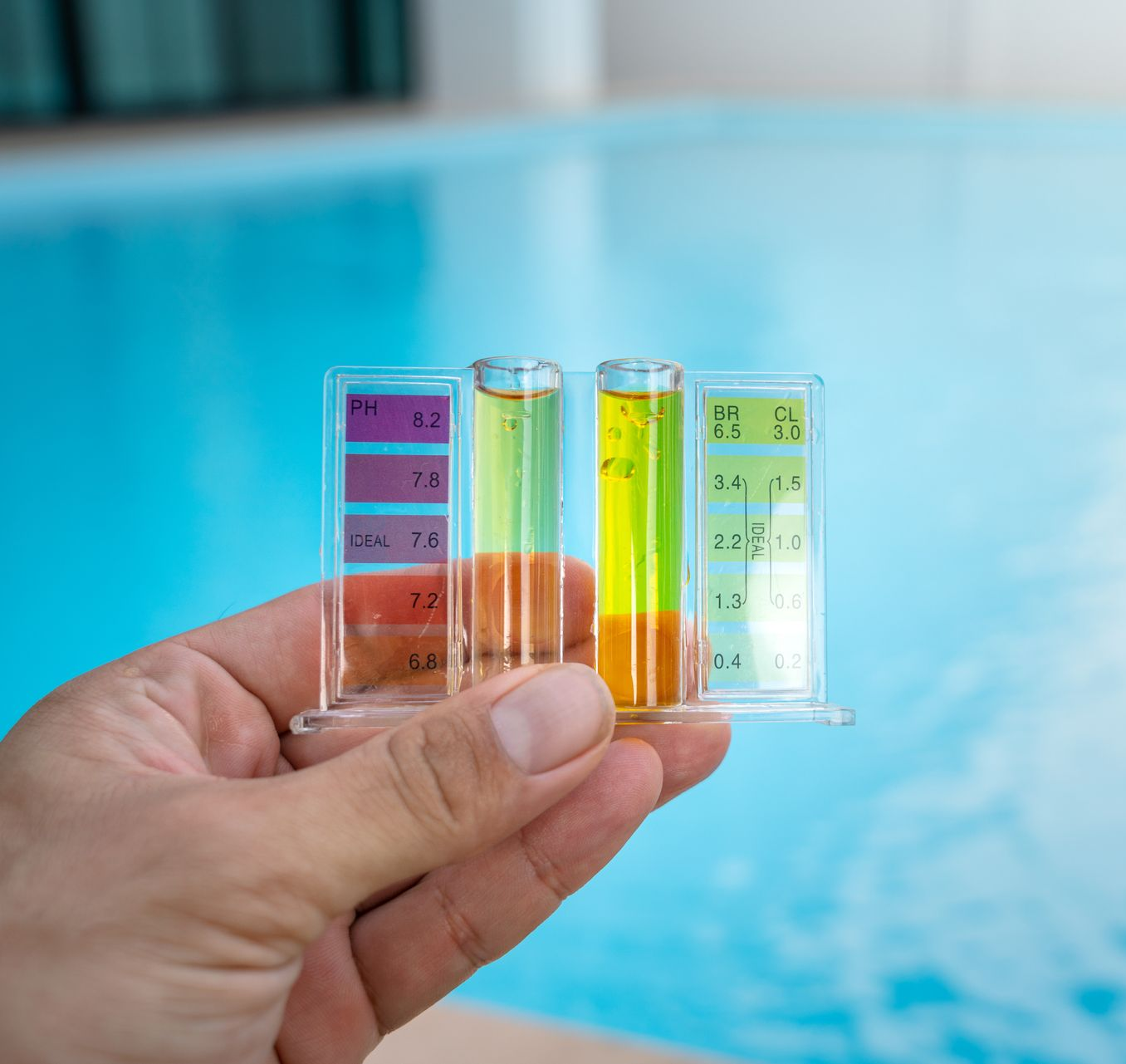 Hand holding pool water test kit, showing pH, chlorine, and bromine levels against a blue pool.