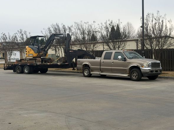 Tan pickup truck towing a trailer with a yellow excavator on an asphalt lot.